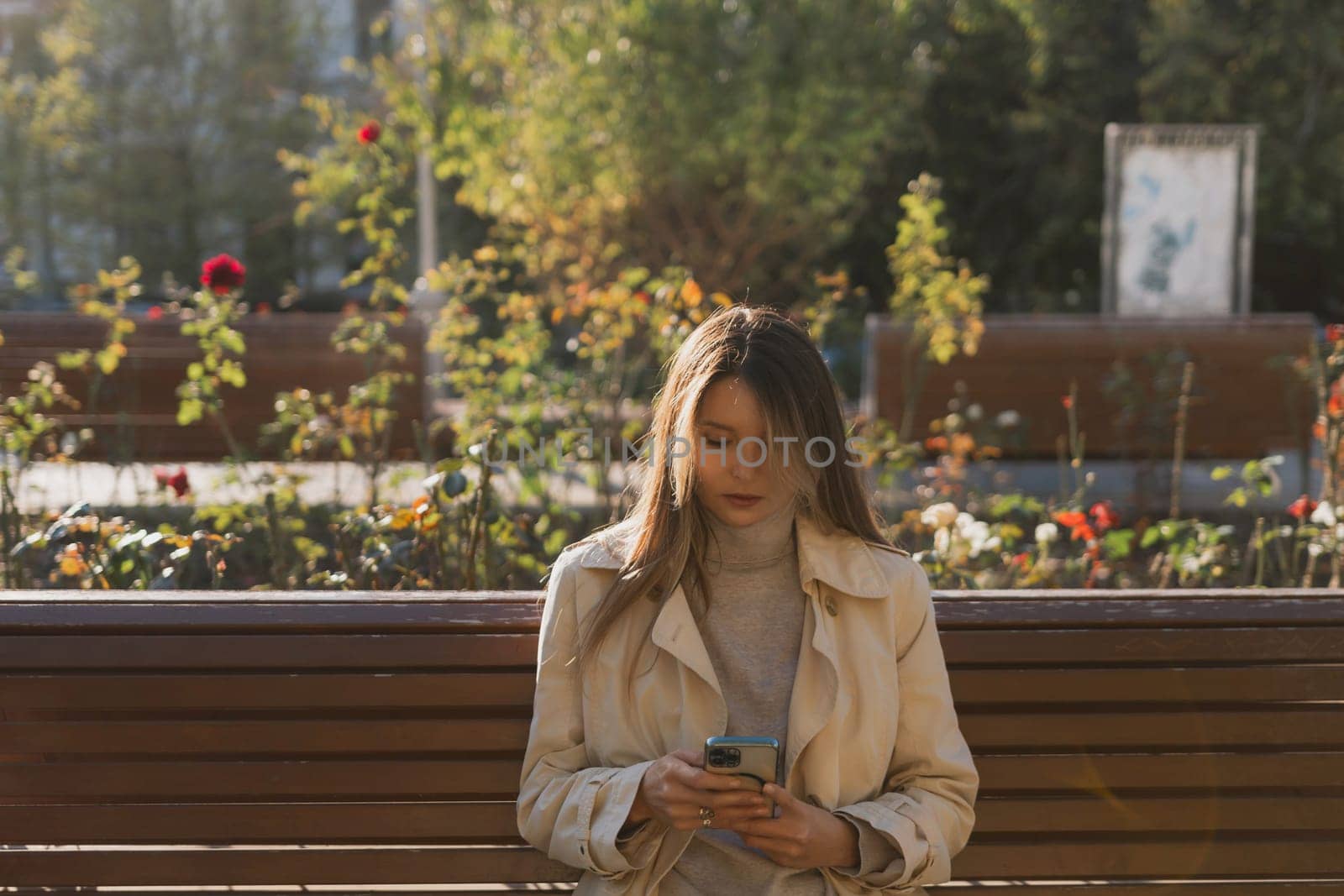 A woman is sitting on a bench and looking at her cell phone. She is wearing a tan coat and she is focused on her phone