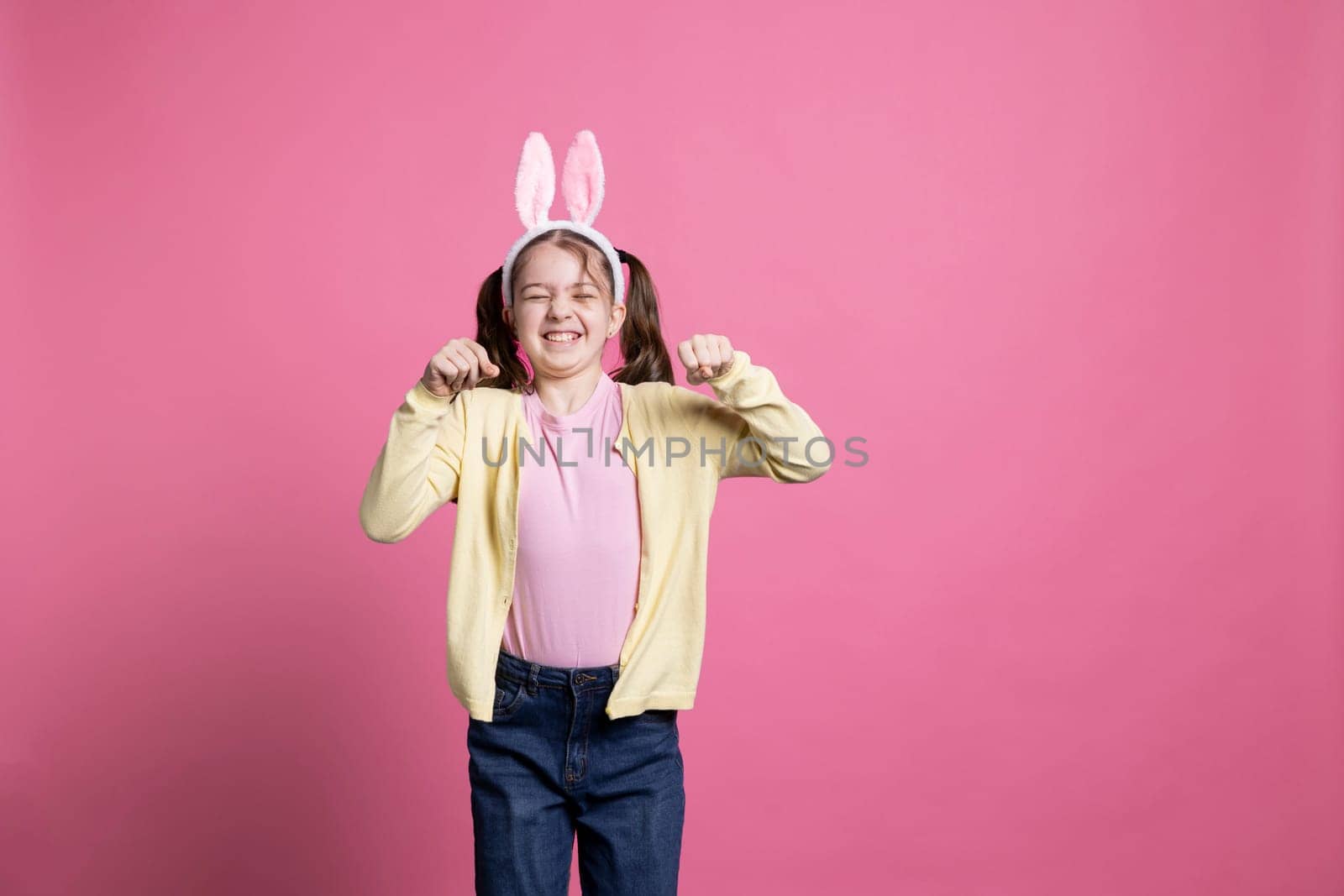 Delightful child imitates a rabbit for the Easter celebration, hopping in the studio while she is wearing bunny ears. Cheerful young kid jumping against pink backdrop, acting goofy and amusing.