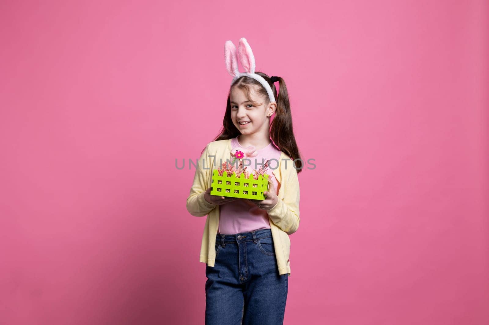 Happy confident girl with bunny ears presenting a basket filled with painted handmade easter decorations over pink background. Young cheerful kid showing festive colorful ornaments in studio.