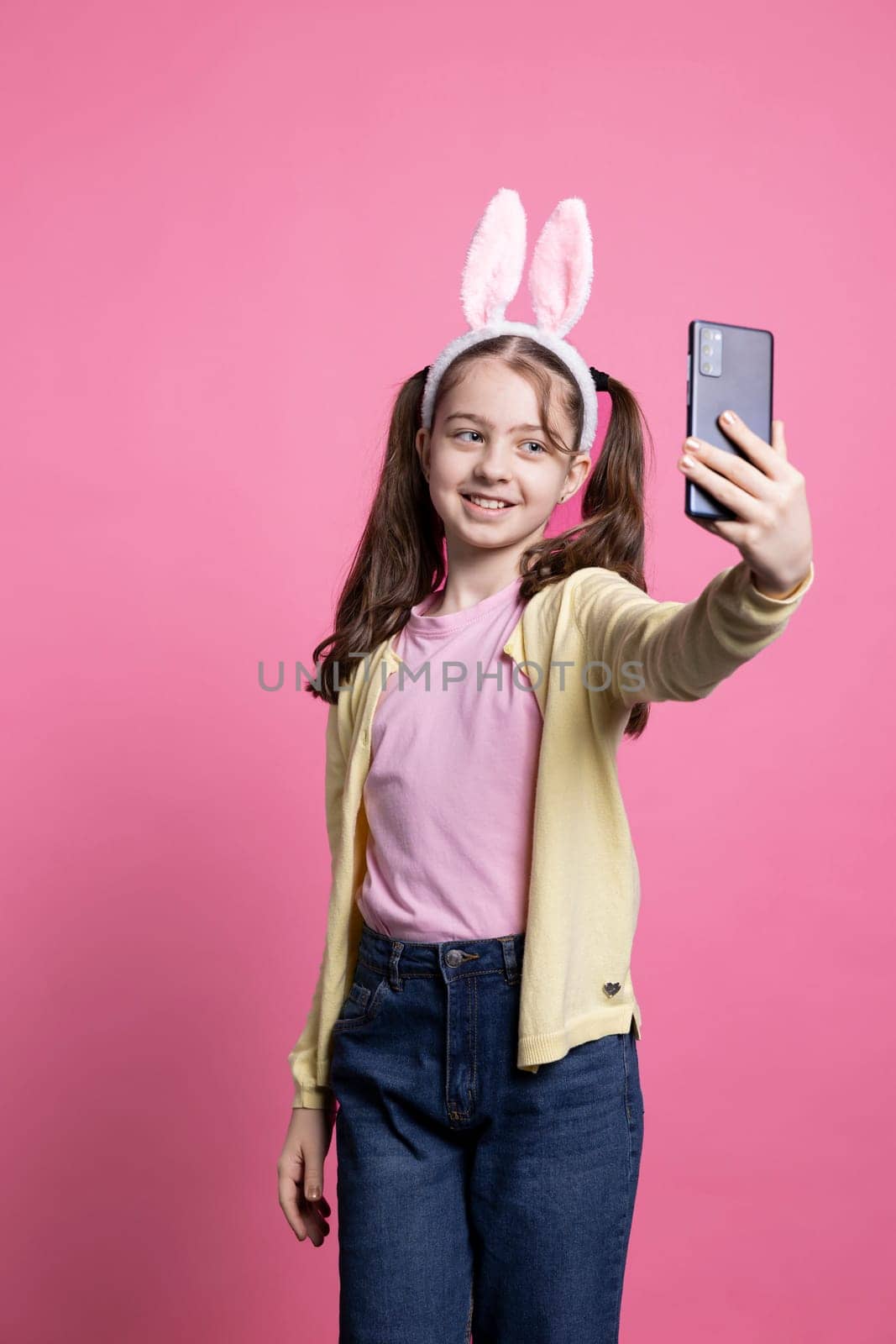 Young excited little kid taking photographs with her phone on camera, smiling in studio and posing against pink background. Positive joyful child takes pictures for easter holiday event.