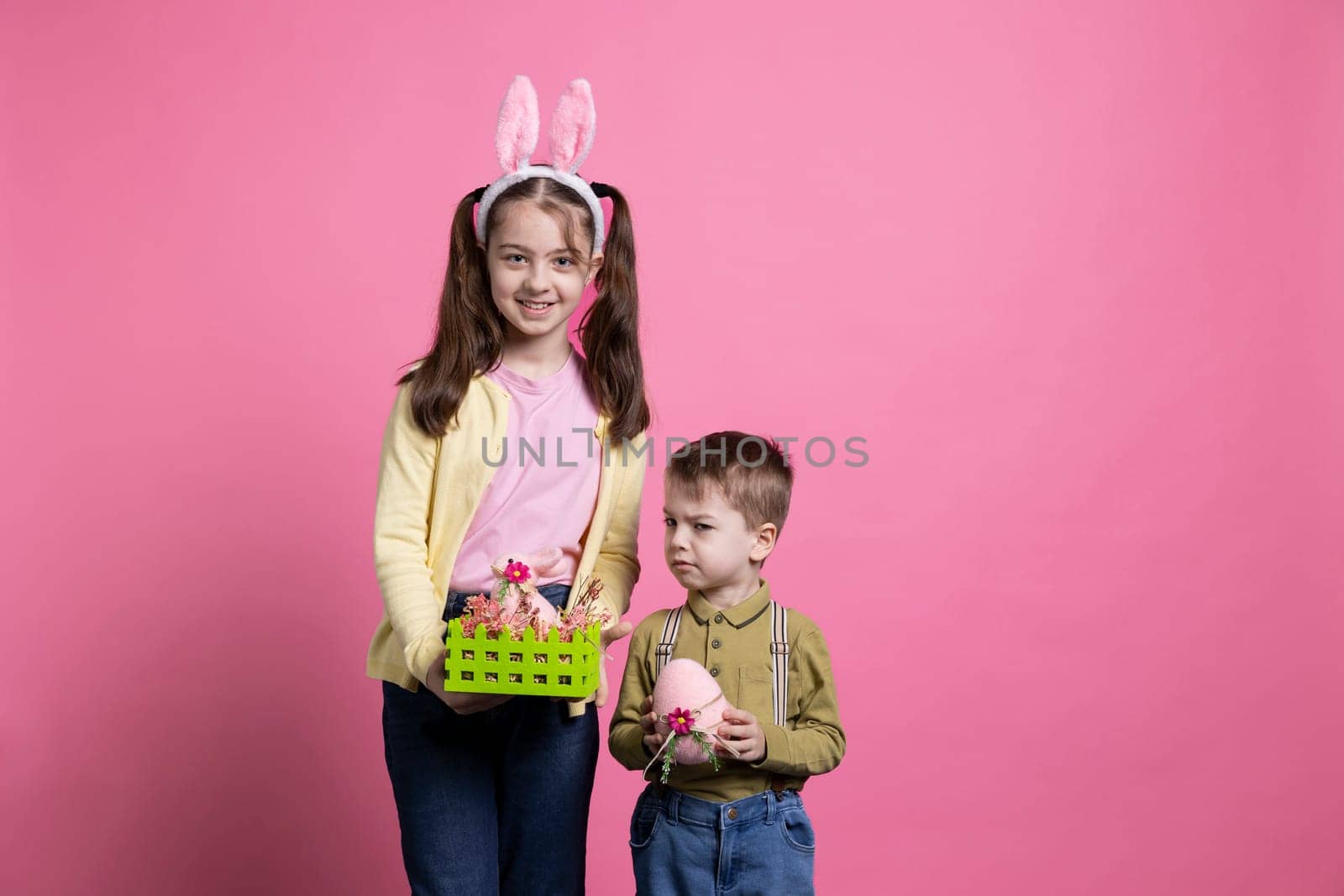 Playful children showing painted eggs and decorating for easter celebration festivity, standing together over pink background. Brother and sister feeling cheerful and enthusiastic about april event.