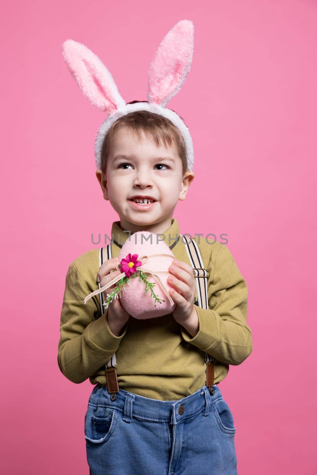 Joyful young child showing a pink egg decorated for easter, feeling happy and excited about spring holiday event. Little cute kid holding handmade festive ornament, wearing bunny ears in studio.