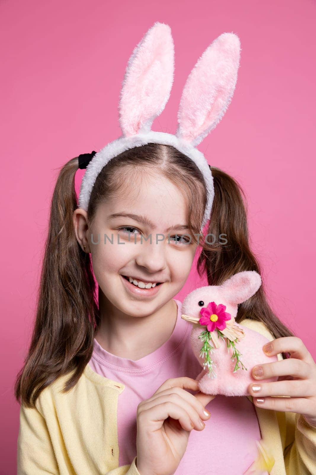 Joyful pretty toddler with bunny ears and pigtails holding a pink rabbit toy, posing with confidence over pink background. Young girl feeling optimistic about easter celebration, colorful decor.