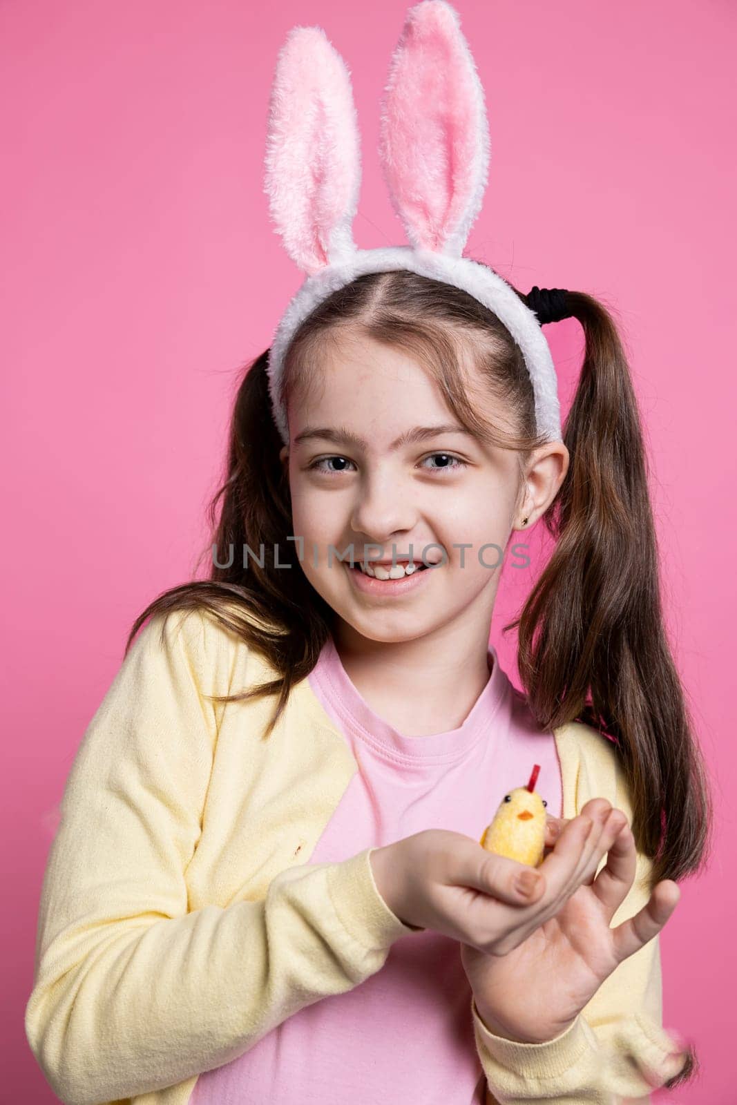Small joyful girl presenting a fluffy chick toy in pink studio, adorable young child with golden stuffed toy over colored background. Little kid with festive bunny ears for seasonal celebration.