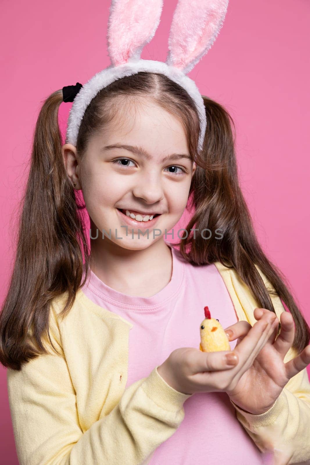 Young cute kid holding a stuffed chick in front of camera, positive excited girl feeling happy about easter celebrations. Small child with fluffy bunny ears smiling over pink background.