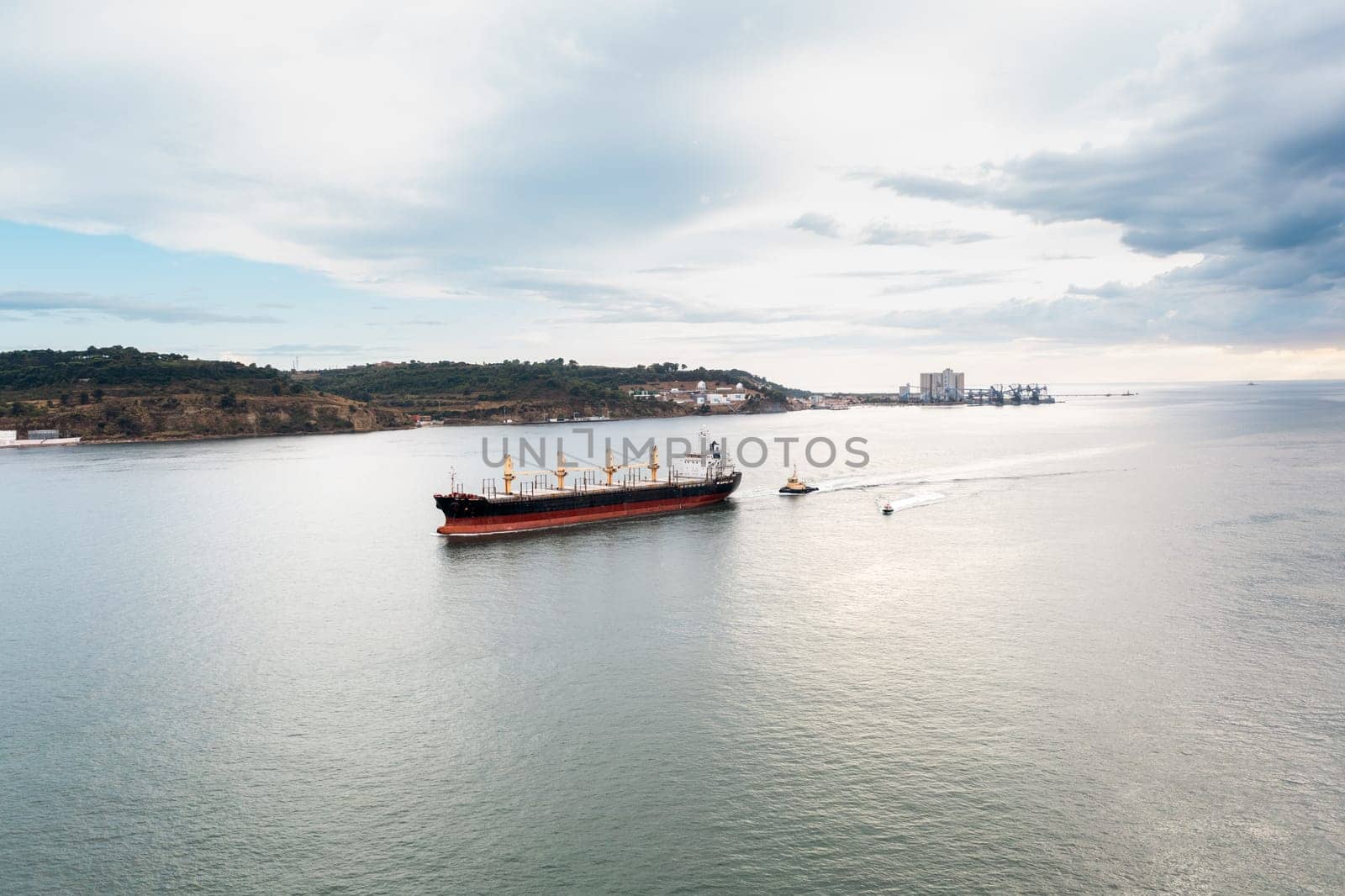 Aerial drone shot of cargo ship and small boat moving in calm sea along mountains under cloudy sky, import export around the world