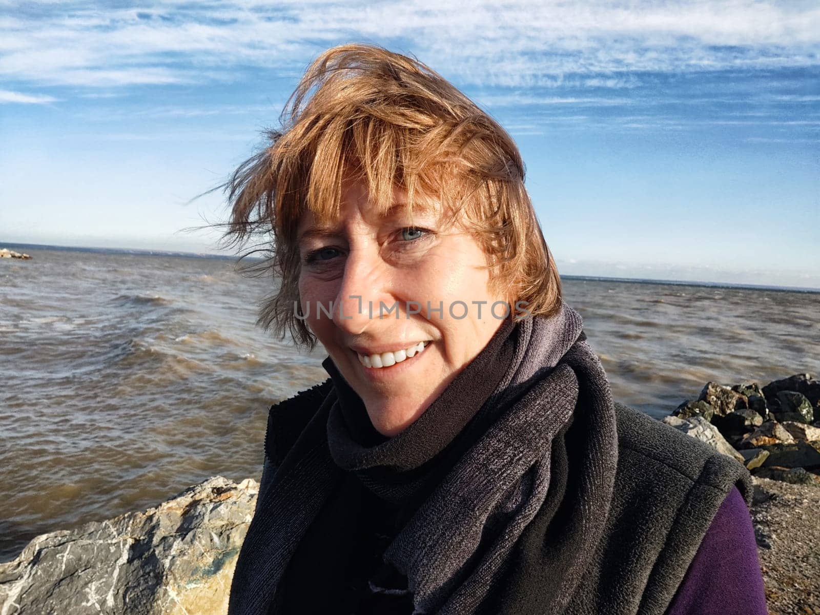 Happy cheerful middle aged woman in scarf taking selfie on nature outdoors and sun with water and waves of sea on the background