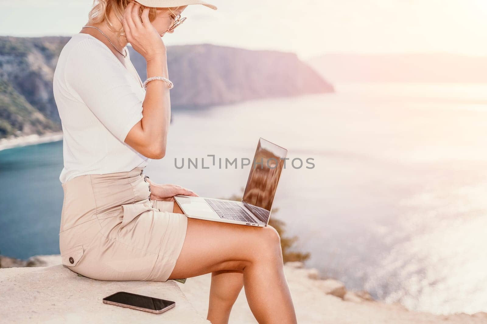 Successful business woman in yellow hat working on laptop by the sea. Pretty lady typing on computer at summer day outdoors. Freelance, travel and holidays concept.