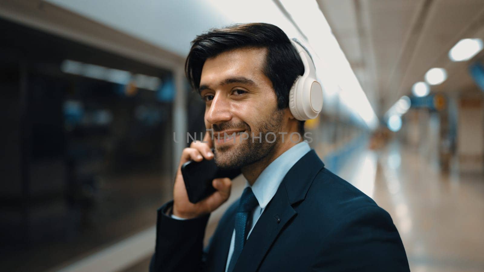 Smart business man calling phone while wearing headphone at train station. Professional executive manager talking to colleague about marketing strategy while waiting for train or subway. Exultant.