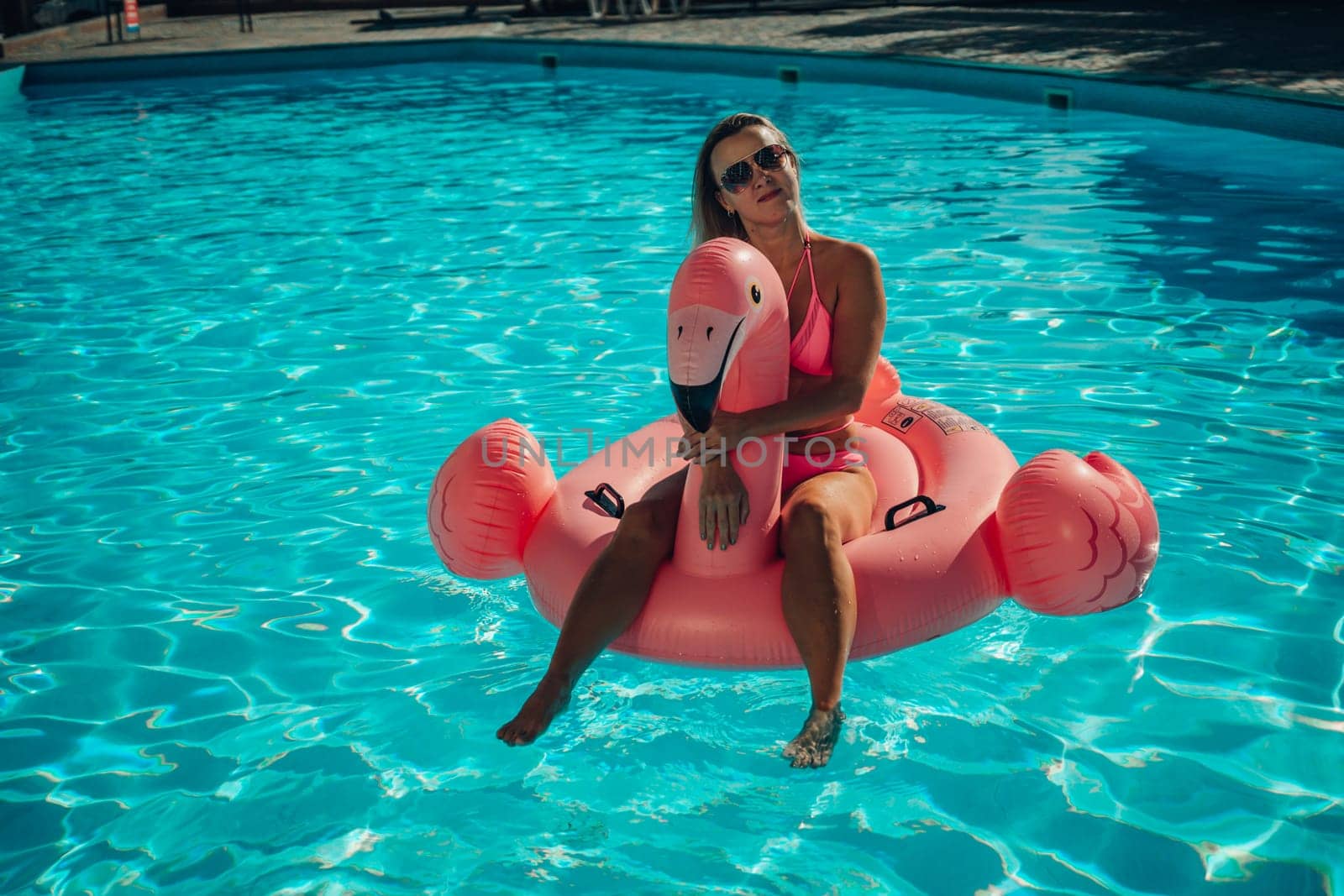 A woman is sitting on a pink inflatable flamingo in a pool. Concept of fun and relaxation, as the woman is enjoying her time in the water by Matiunina