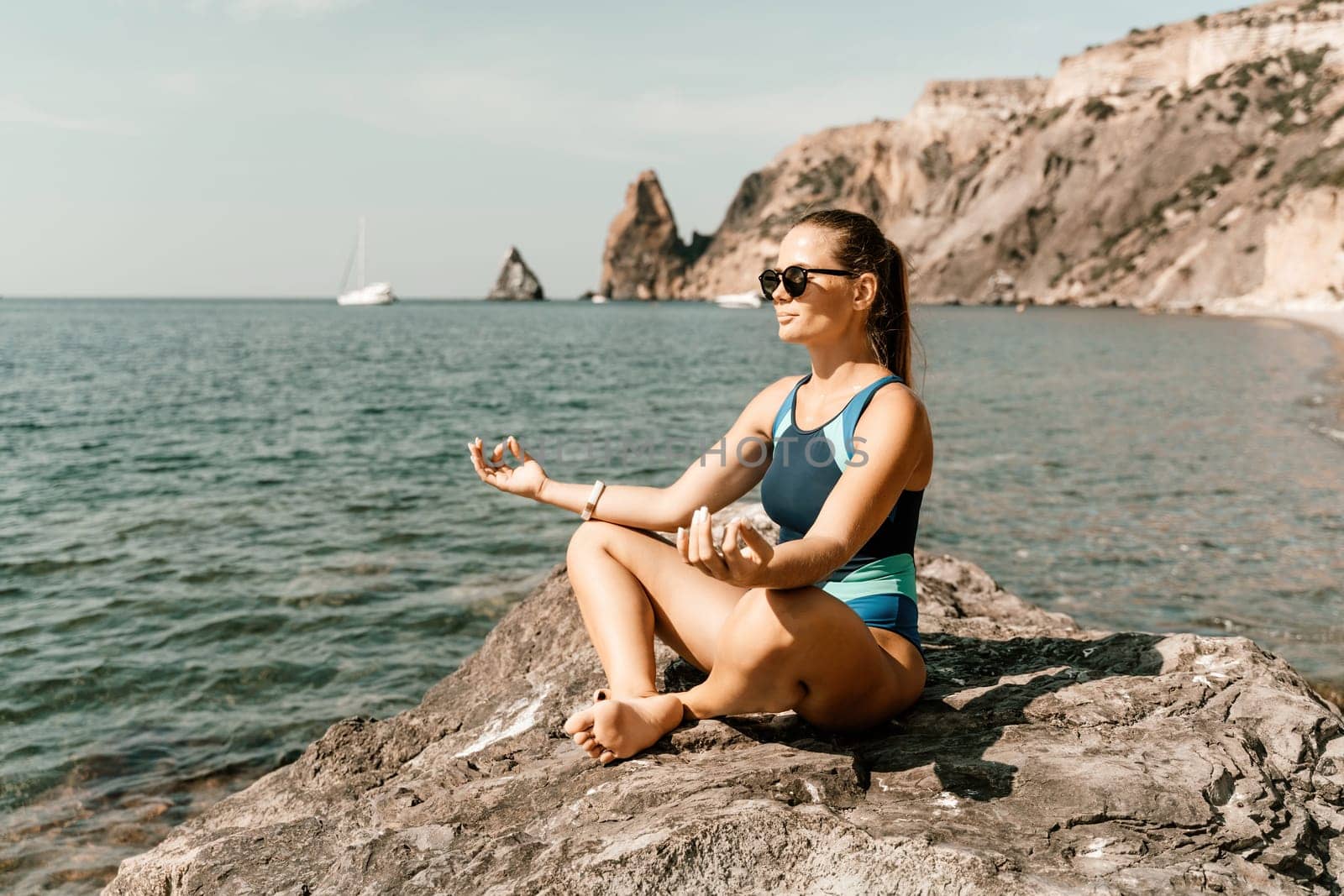 Yoga on the beach. A happy woman meditating in a yoga pose on the beach, surrounded by the ocean and rock mountains, promoting a healthy lifestyle outdoors in nature, and inspiring fitness concept