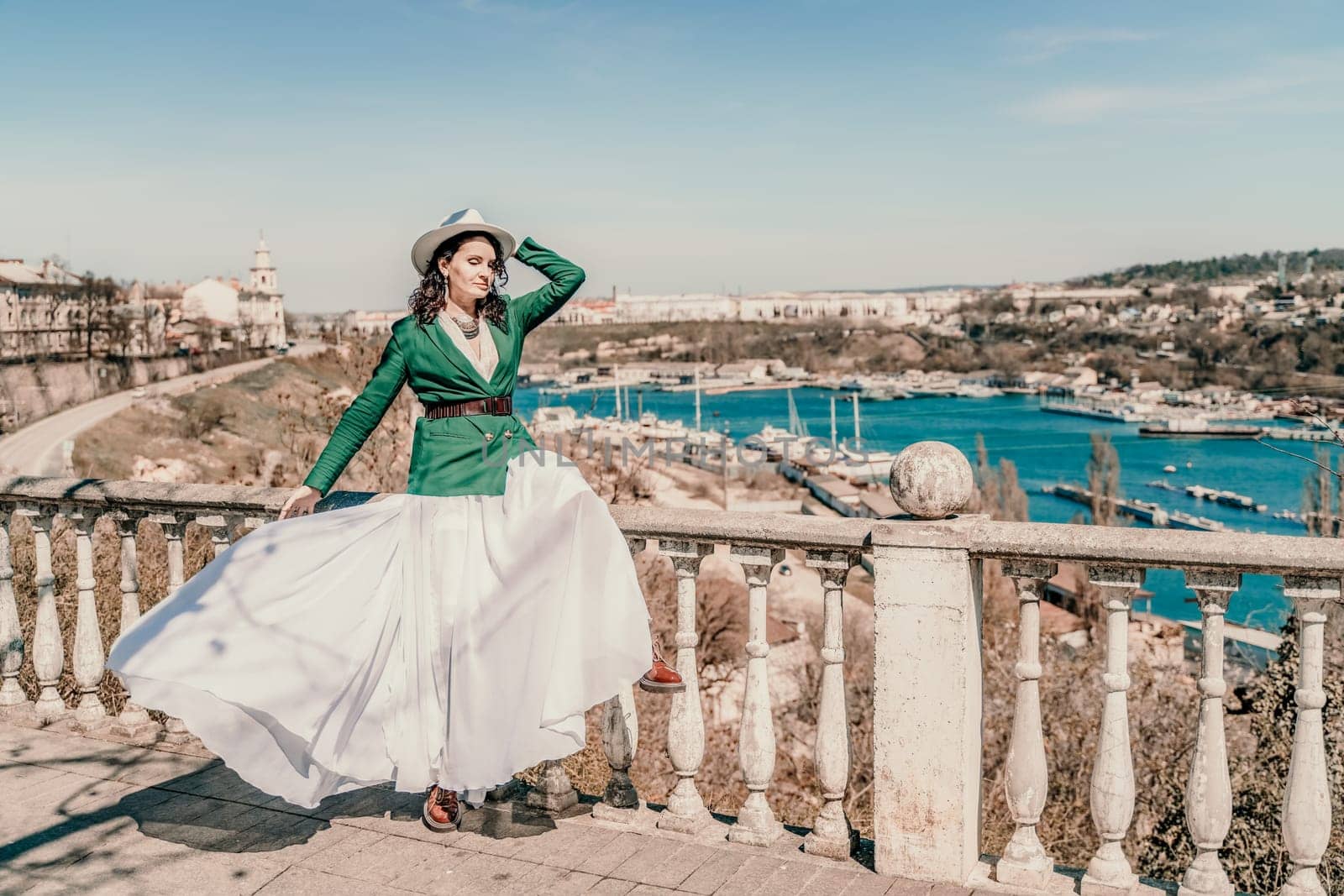 Woman walks around the city, lifestyle. A young beautiful woman in a green jacket, white skirt and hat is sitting on a white fence with balusters overlooking the sea bay and the city. by Matiunina