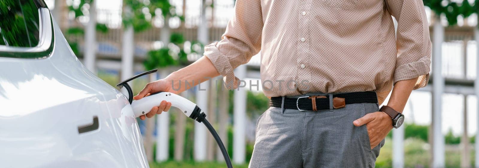 Young man recharge electric car's battery from charging station in city commercial parking lot. Rechargeable EV car for sustainable environmental friendly urban travel. Panorama Expedient