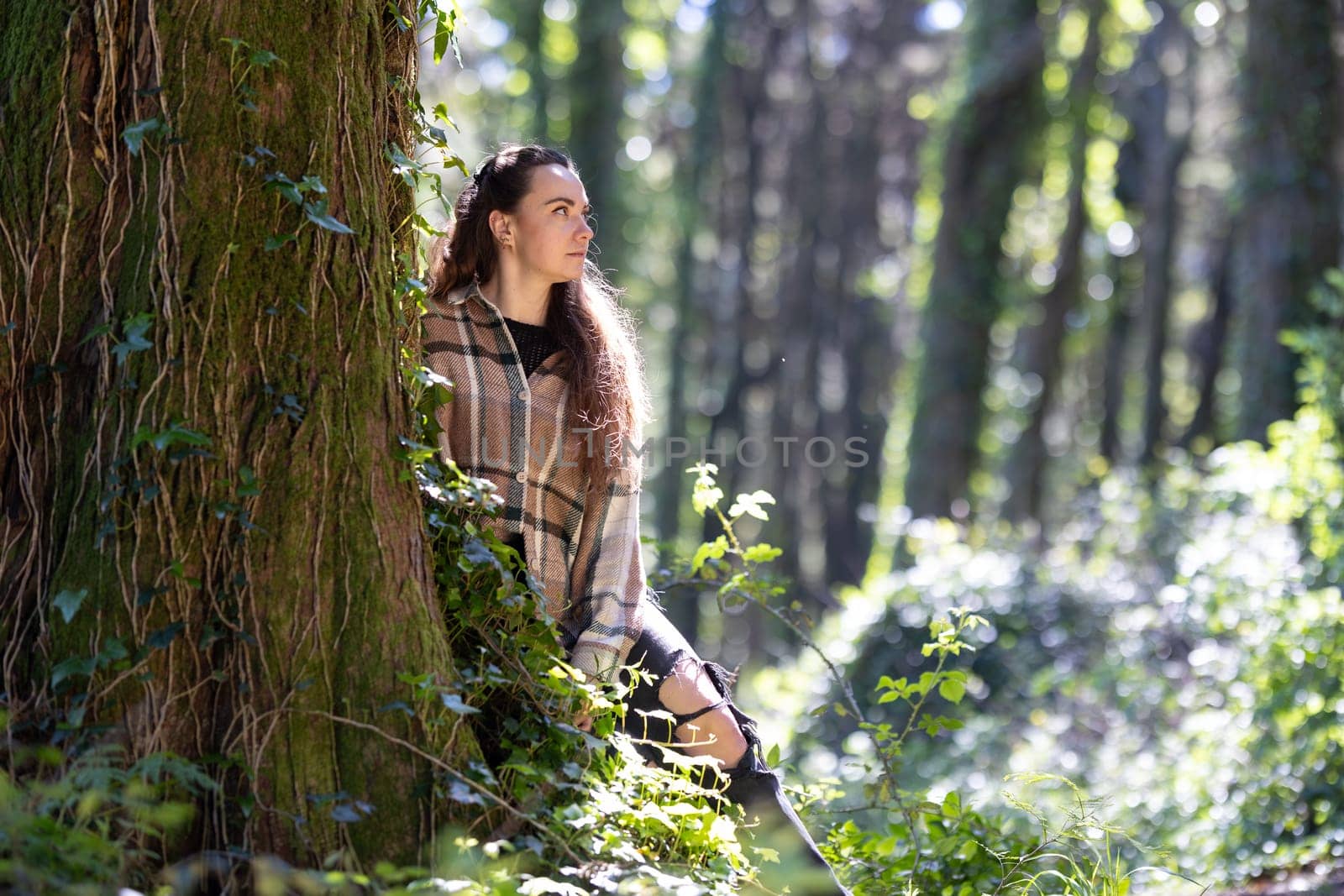 A woman stands next to a tree, surrounded by a forest. She appears to be enjoying the outdoors with friends on a sunny day.