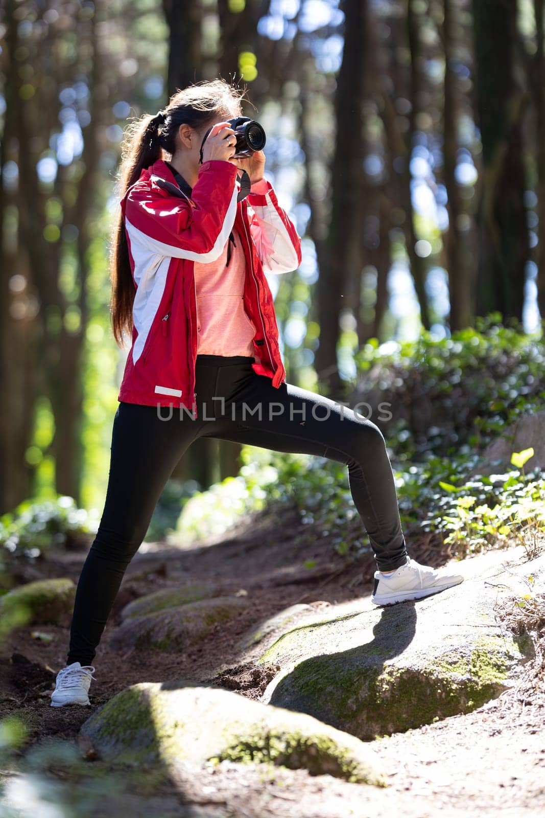 A woman stands in the woods, holding up her phone to take a selfie. The trees and greenery of the forest surround her as she captures the moment.