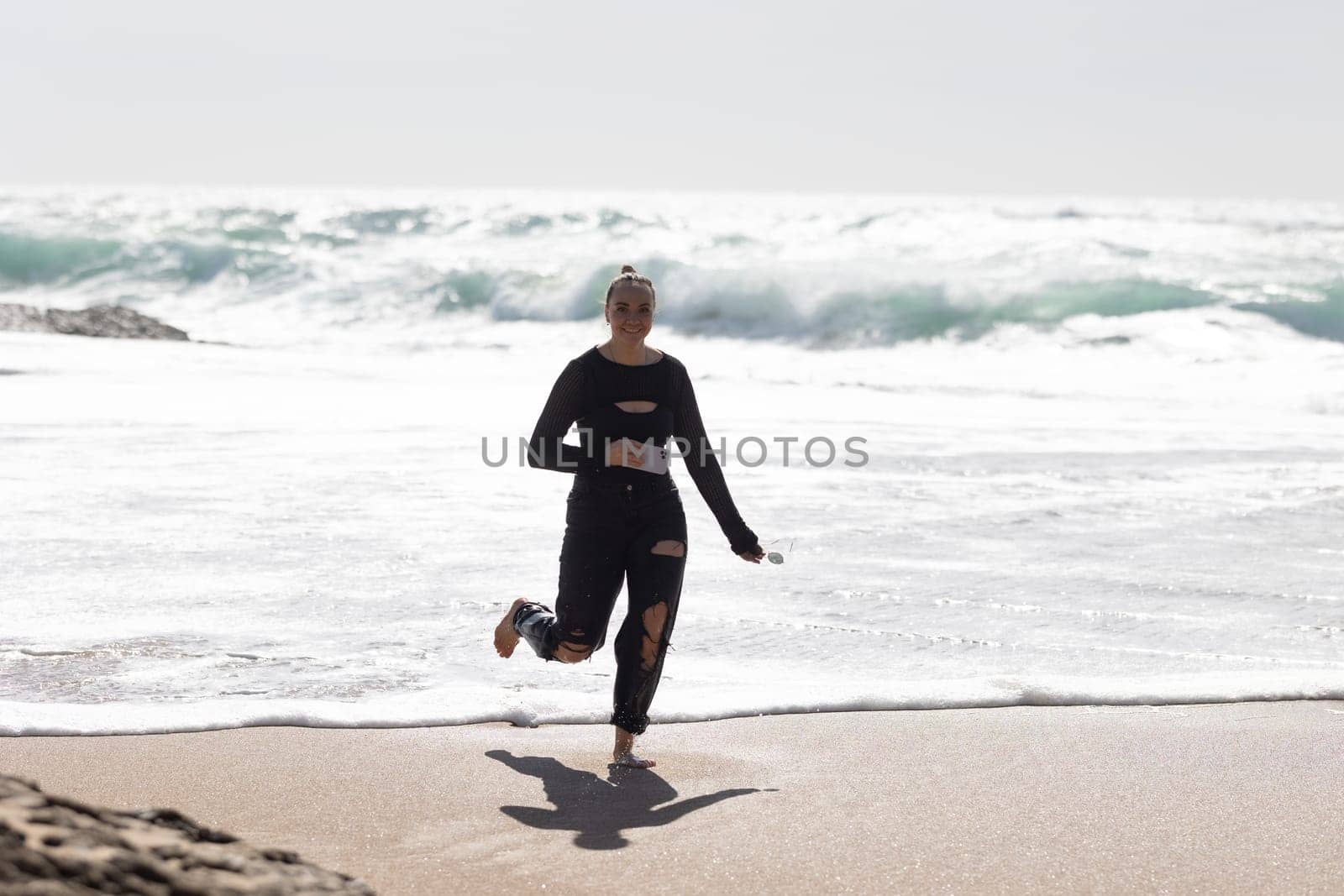 A woman wearing a wet suit is running on the beach. Her hair is flying behind her as she moves energetically along the sandy shore with friends.