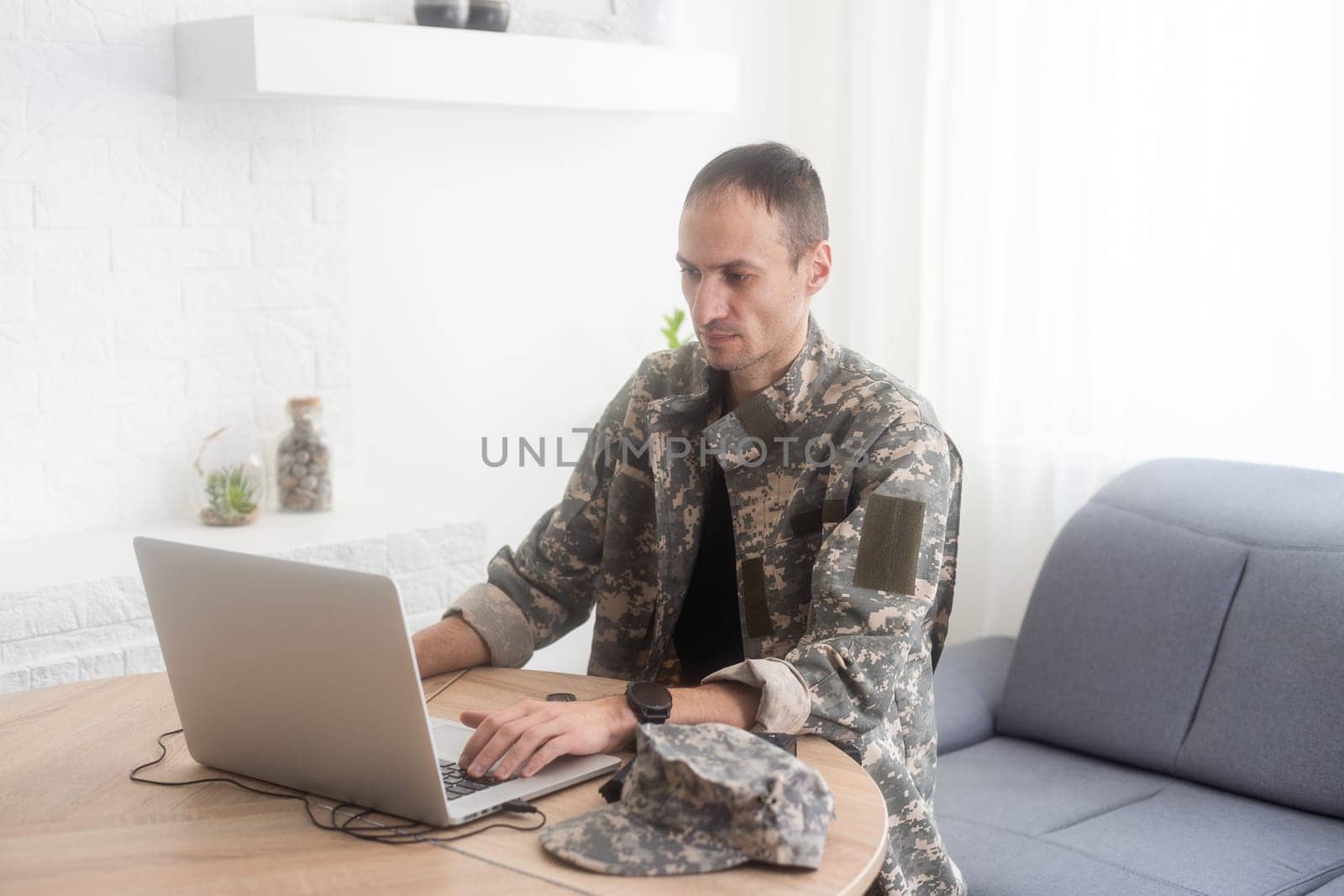 Soldier in office. Serious young military man typing on laptop while working in headquarters building. Caucasian man in camouflage uniform is sitting in bright room at table with notebook and cap. by Andelov13