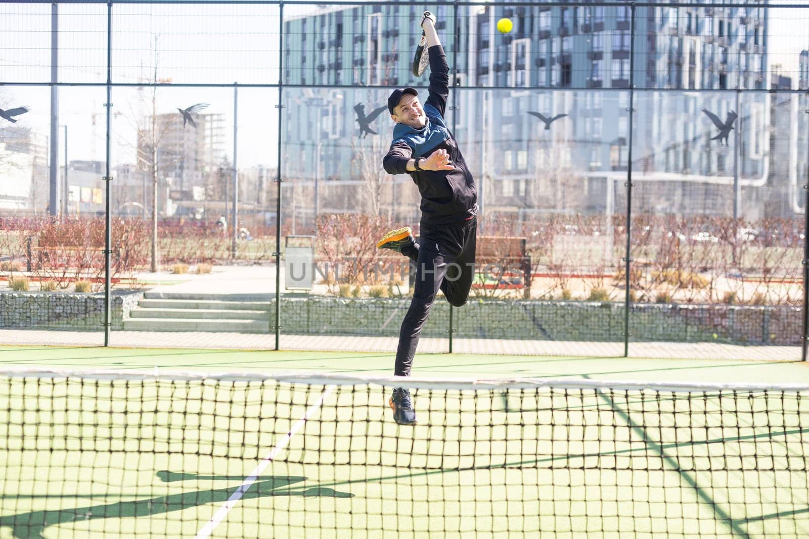 A padel player jump to the ball, good looking for posts and poster. Man with black racket playing a match in the open behind the net court outdoors. Professional sport concept with space for text by Andelov13