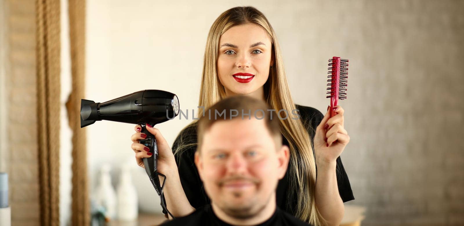 Blonde Hairstylist Using Dryer and Comb for Man.Young Hairdresser Styling Male Haircut with Hairdryer and Hairbrush. Woman Stylist Holding Tool for Hairstyling. Beautician and Client Looking at Camera