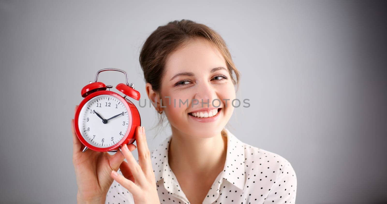 Adult smiling caucasian woman hold red alarm clock on gray background.