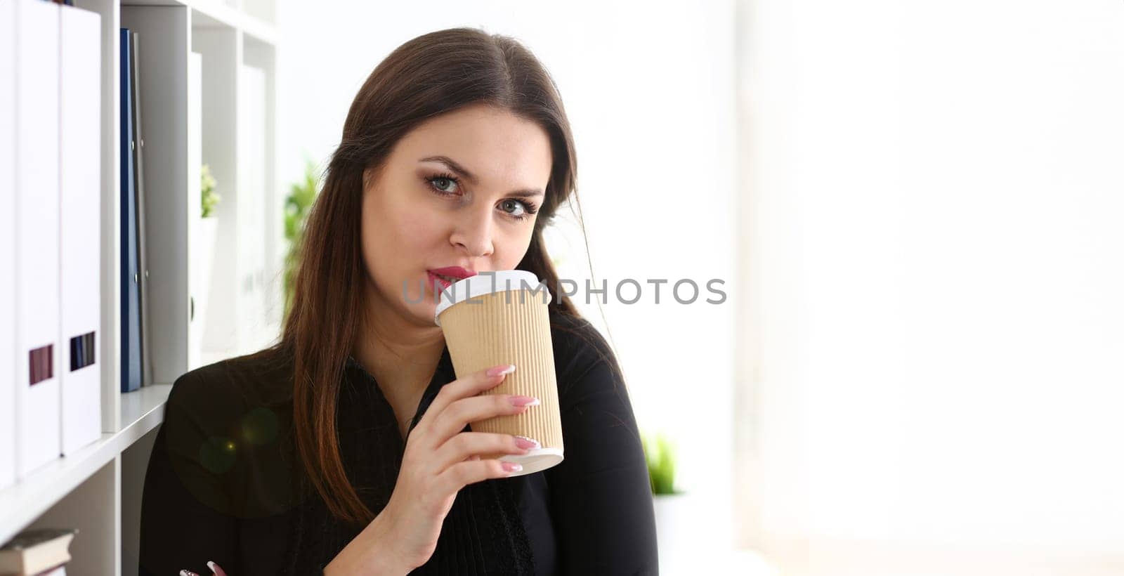 Businesswoman at workplace in office portrait during break drinks coffee from white cup smiling and looking at camera creates visibility of work and success causes trust.