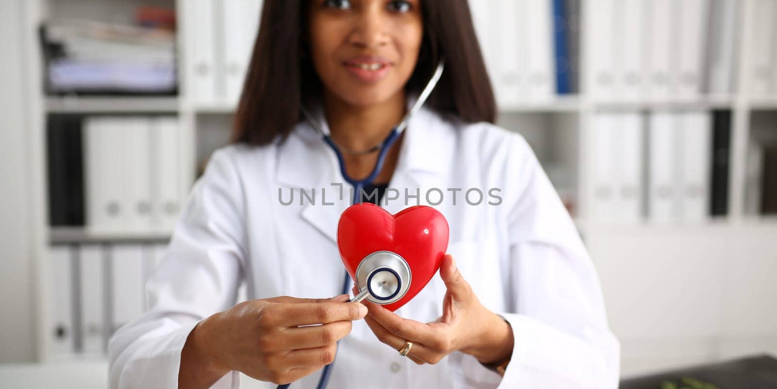 Beautiful black smiling female doctor hold in arms red toy heart closeup. Cardio therapeutist student education CPR 911 life save physician make cardiac physical pulse rate measure arrhythmia