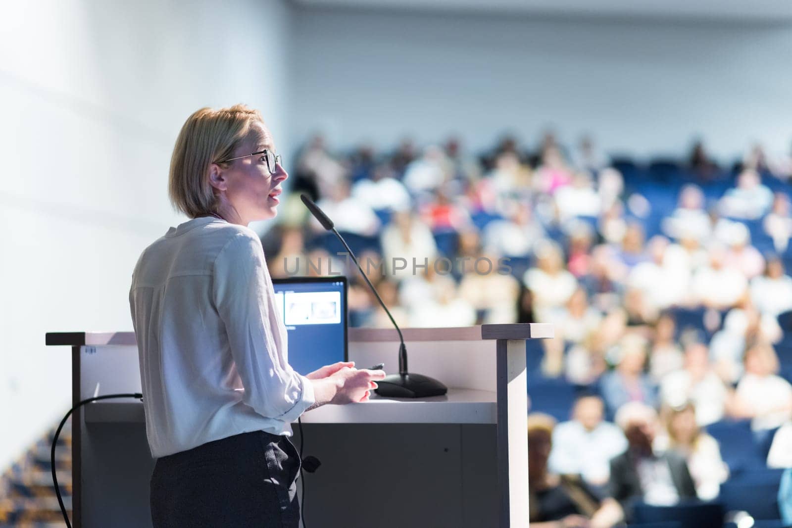 Female speaker giving a talk on corporate business conference. Unrecognizable people in audience at conference hall. Business and Entrepreneurship event