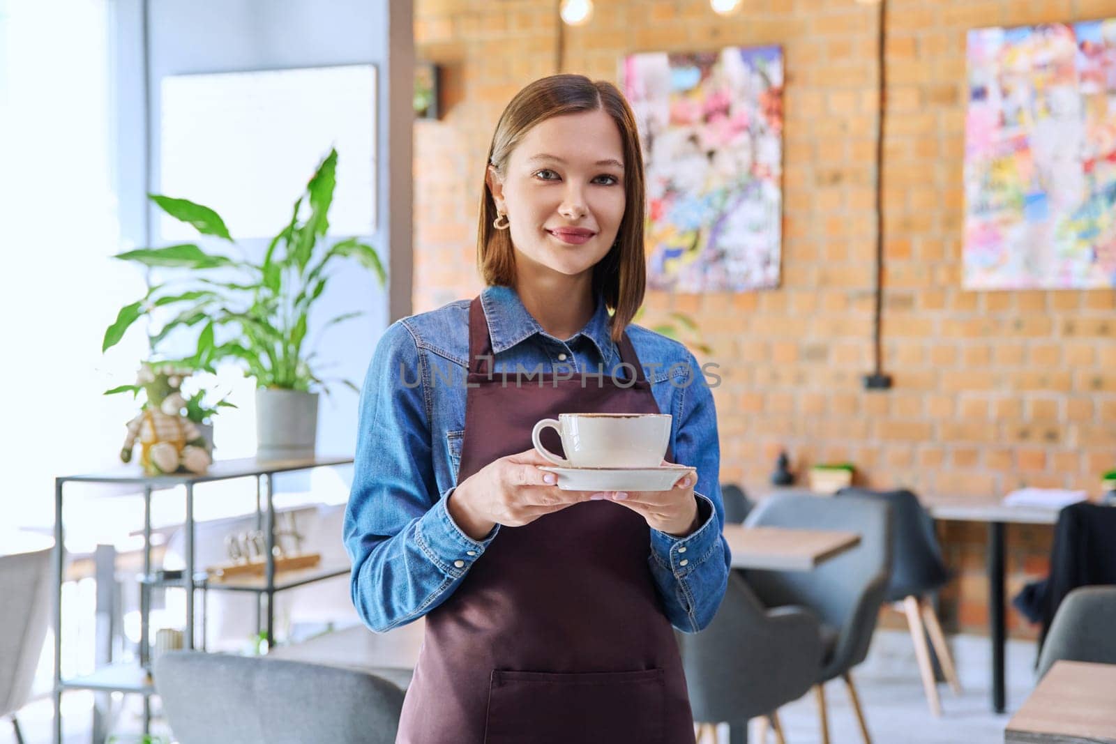 Successful young woman service worker owner in apron with cup of coffee looking at camera in restaurant cafeteria coffee pastry shop interior. Small business staff occupation entrepreneur work