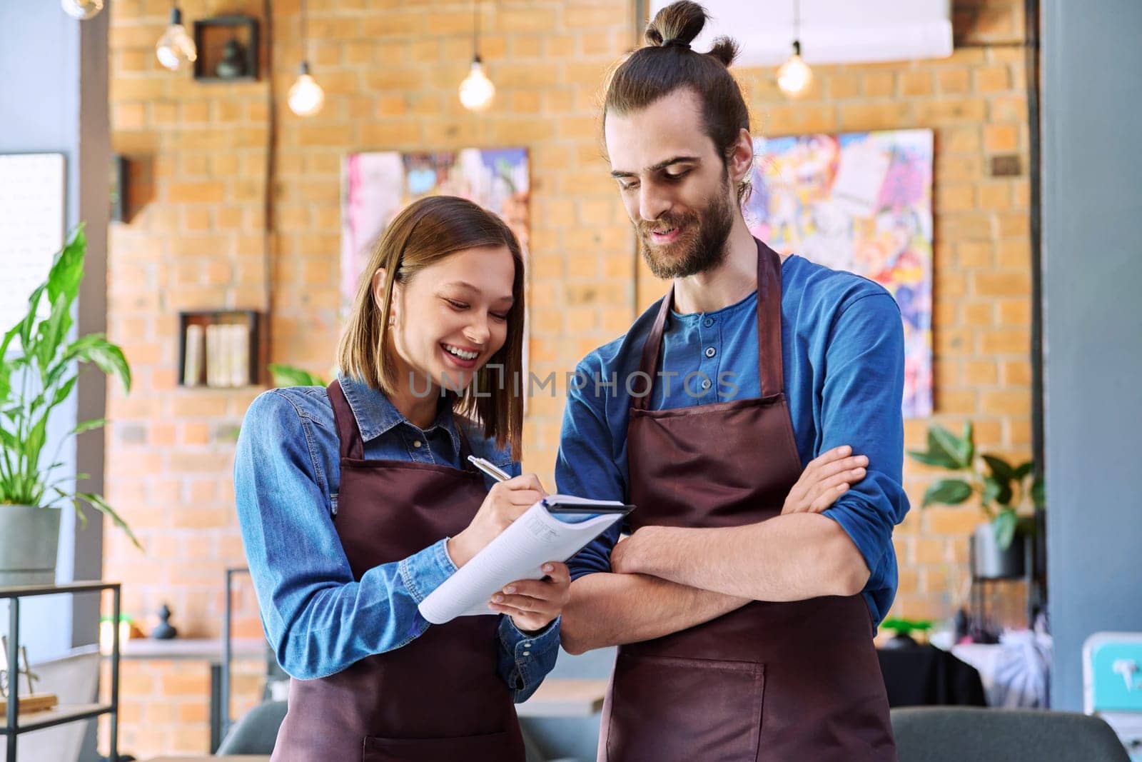 Small business team confident successful colleagues partners owners young man woman in aprons writing at working notebook at workplace in restaurant coffee shop cafeteria. Partnership teamwork work