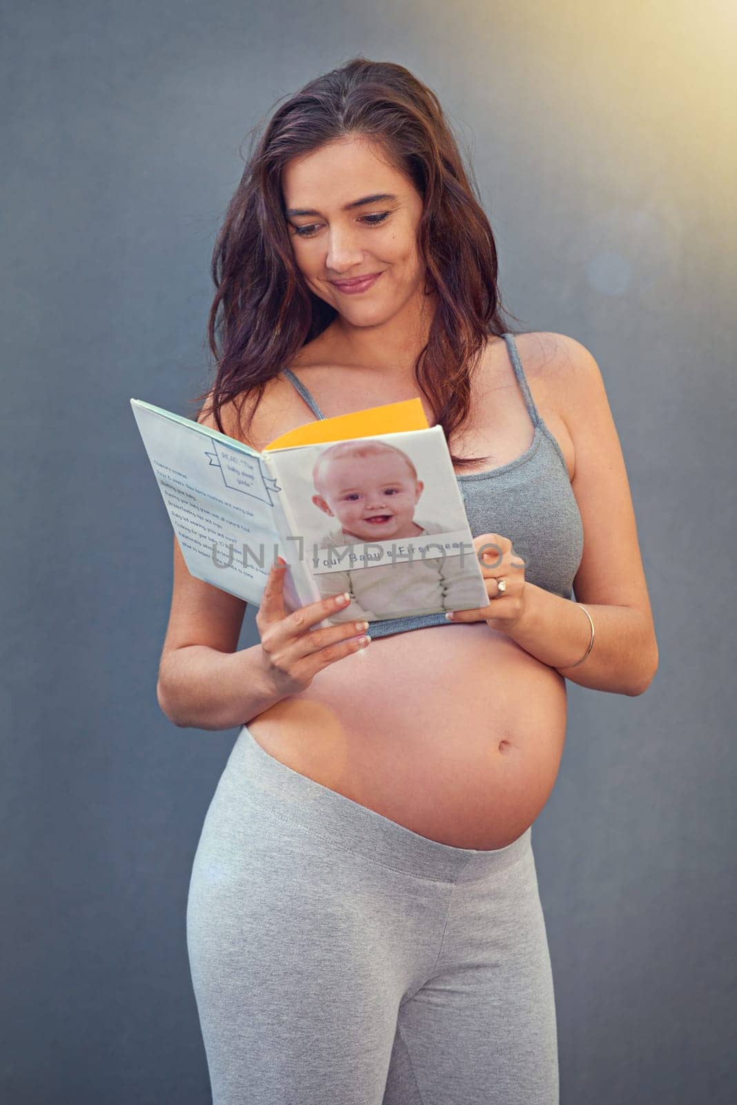 Pregnant woman, baby book and smile in studio for maternity and prenatal wellness for pregnancy and motherhood. Young person, happy and isolated with growing belly and read for childbirth wellbeing.