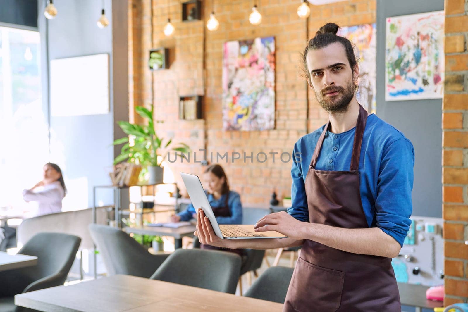 Successful young man service worker owner in apron holding using laptop computer looking at camera in restaurant cafeteria coffee pastry shop interior. Small business staff occupation entrepreneur work