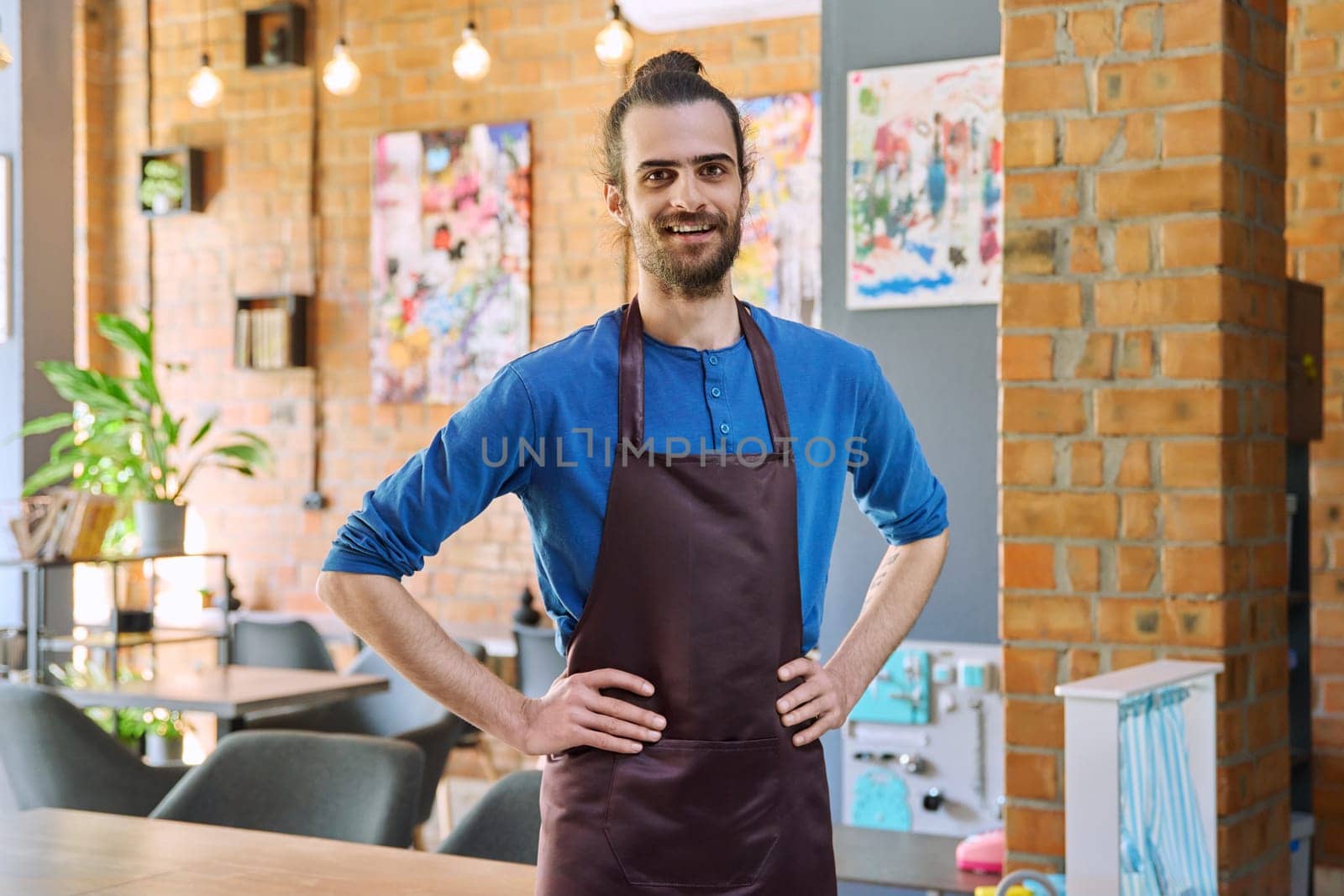 Young man worker, owner in apron looking at camera in restaurant, coffee shop by VH-studio