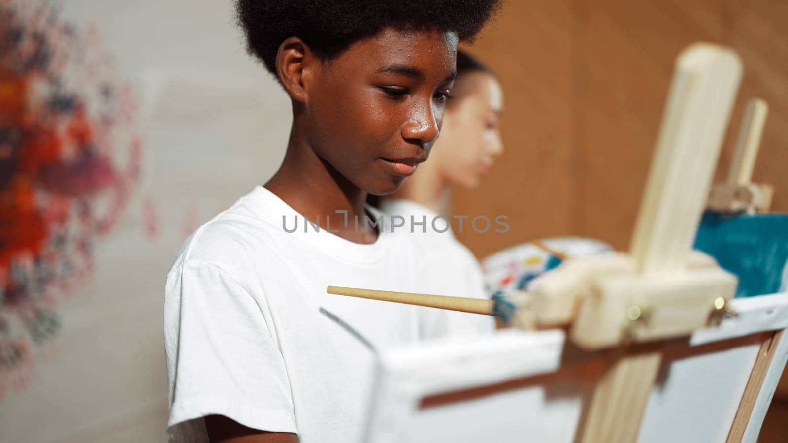 African boy painting canvas with watercolor while happy caucasian girl draw cool tone picture at colorful stained wall. Multicultural highschool student attend creative activity together. Edification.