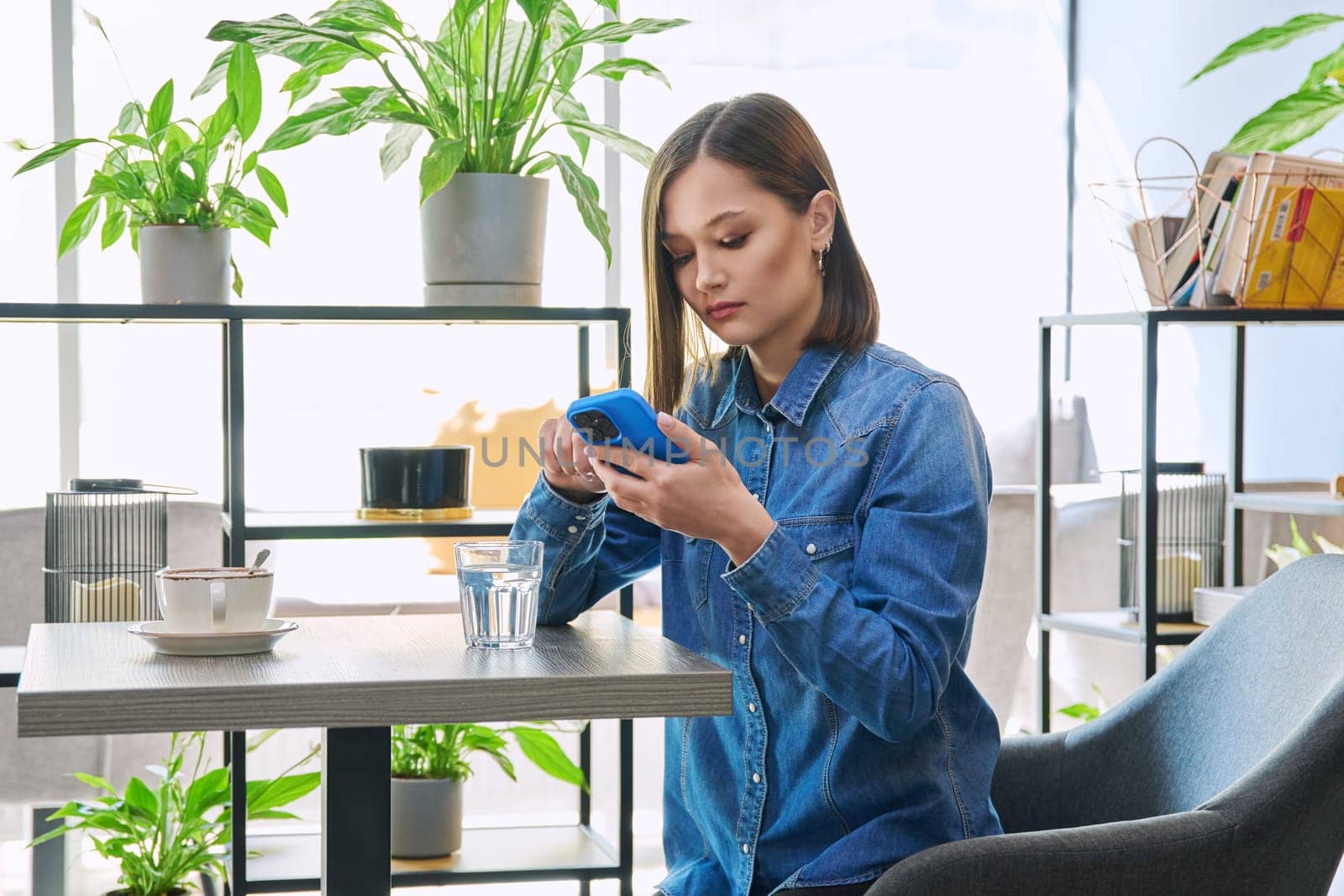 Young attractive woman using smartphone, drinking cup of coffee, glass of water, sitting in cafeteria, cafe. Serious female holding mobile phone, texting messages. Technology lifestyle youth concept