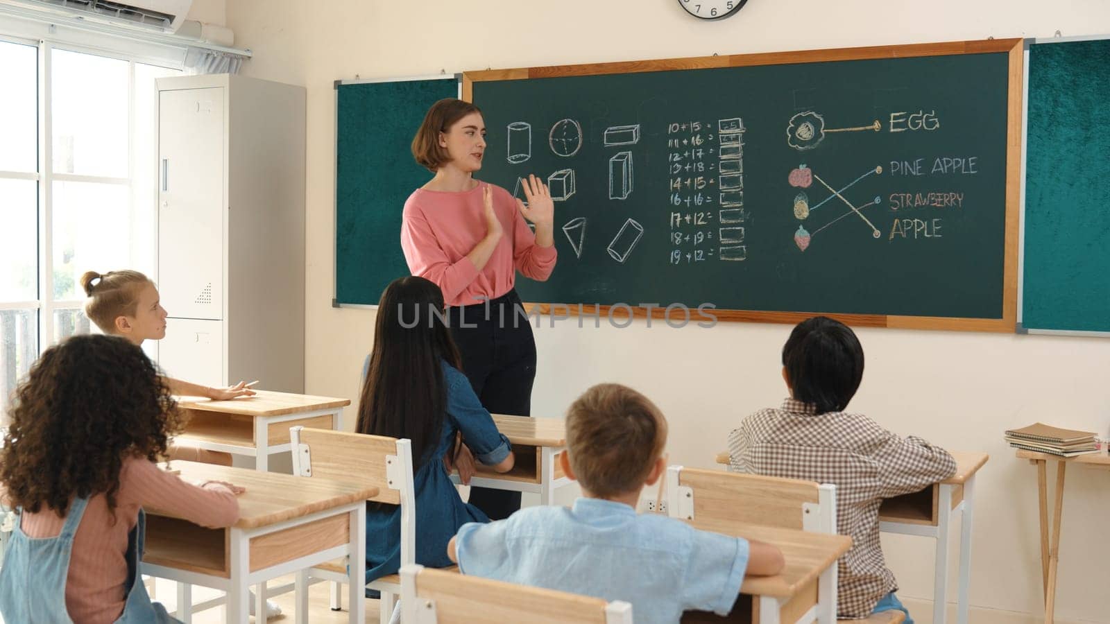 Smart teacher checking cute student homework while explaining idea at class. Attractive instructor walking around classroom while looking at notebook and talking about Math theory to child. Pedagogy.