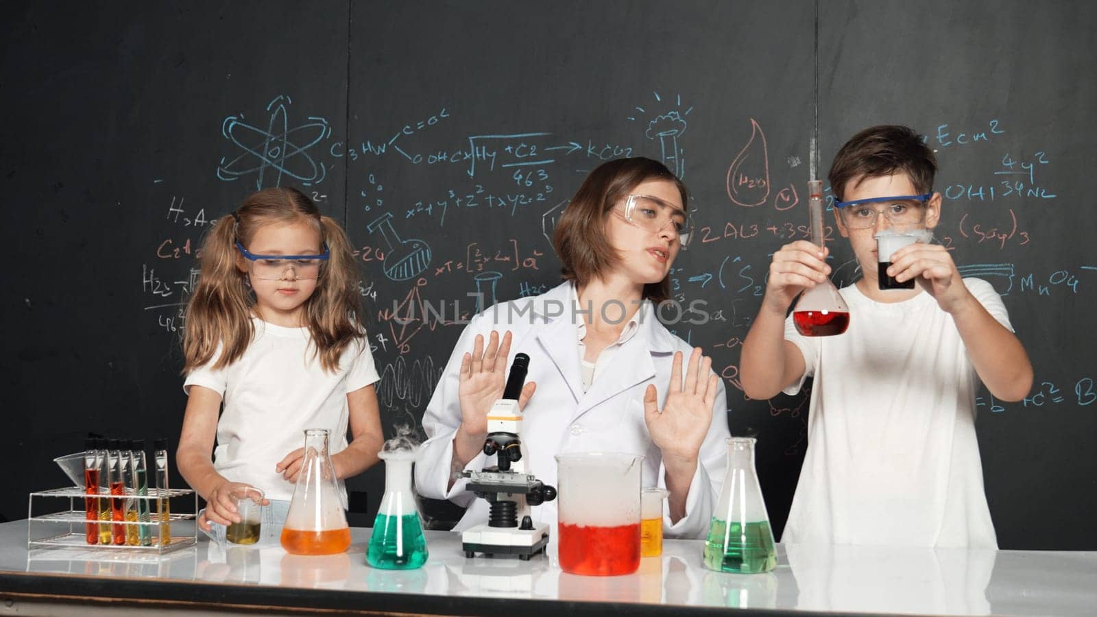 Caucasian boy mixing chemical liquid while teacher giving advice. Professional instructor wearing lab suit looking for diverse student at table with beaker filled with colored solution. Erudition.