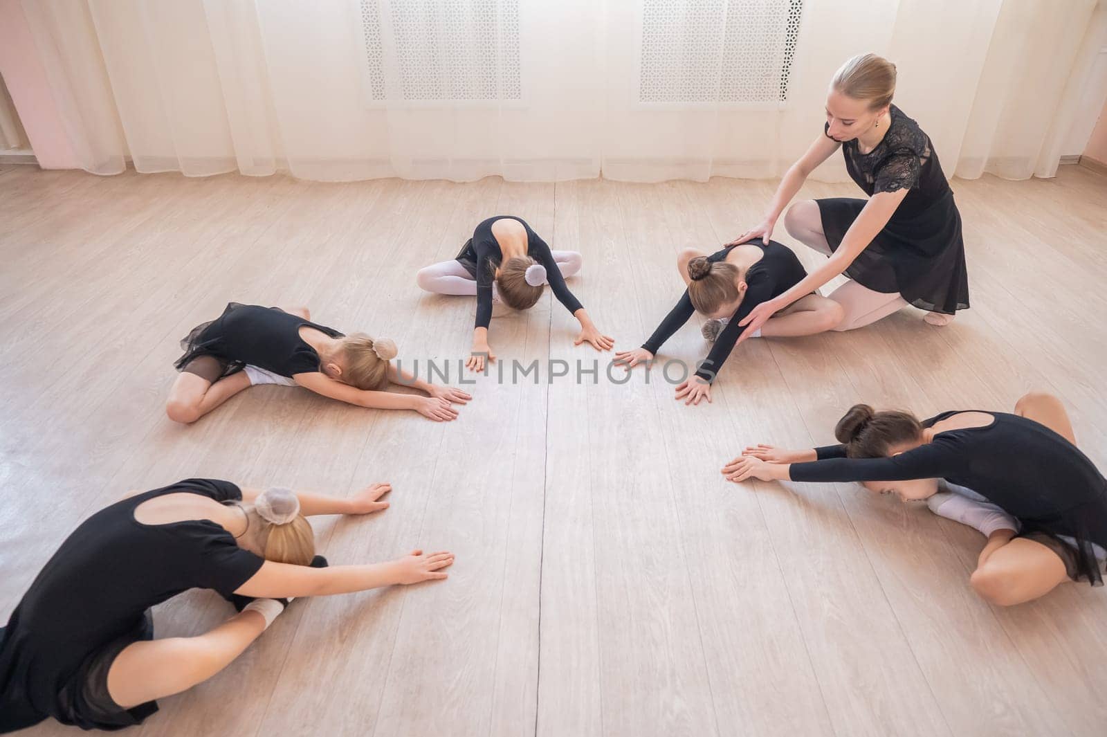 Caucasian woman and five little girls sit in a circle and do stretching at a ballet school