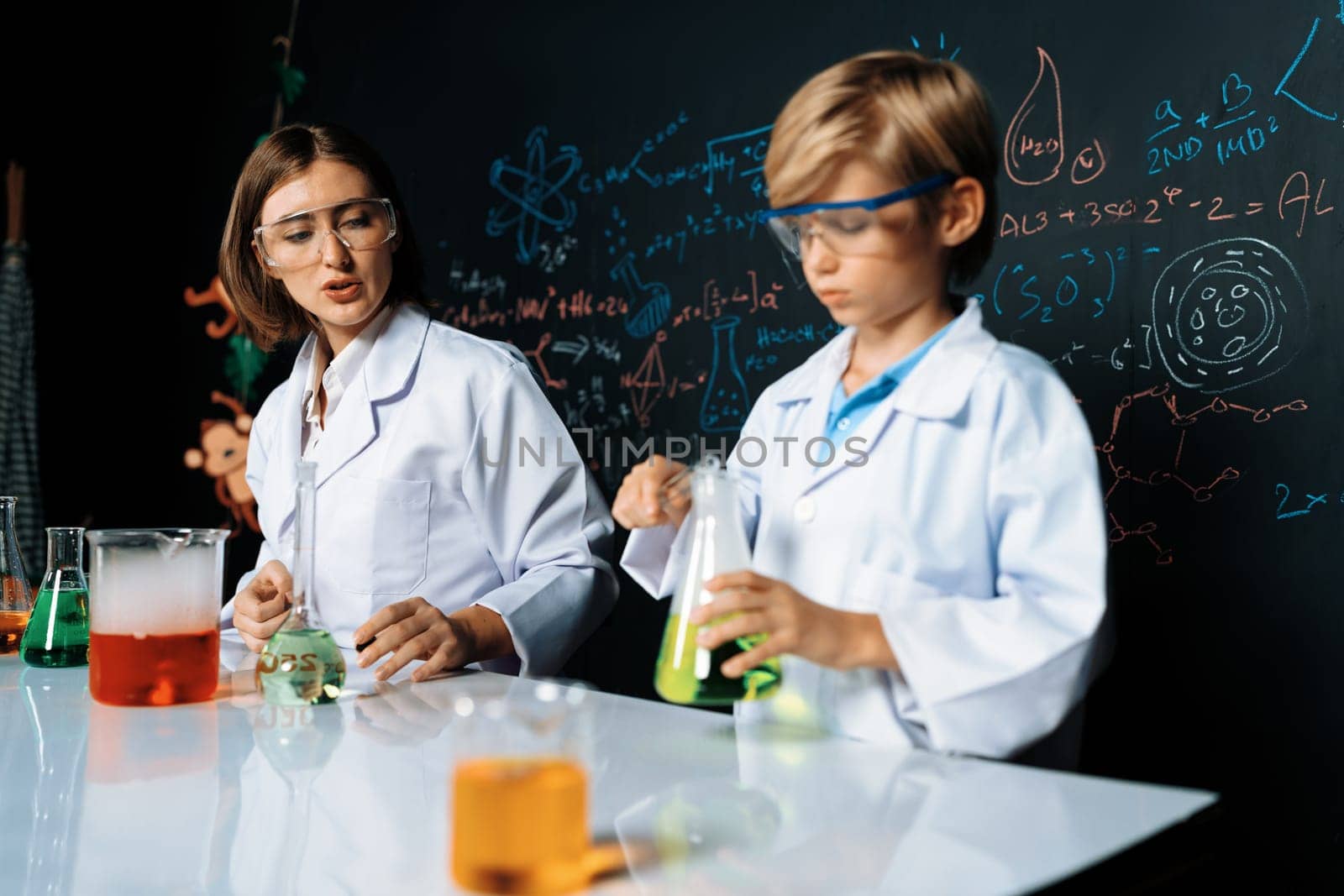 Teacher support schoolboy in laboratory they wear lab coat and glasses stand and experiment about science of chemistry in STEM class. Student tong solid down to yellow liquid in beaker. Erudition.