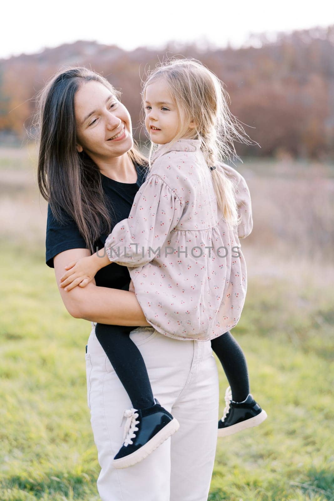 Smiling mother looking at little girl sitting in her arms while standing on the lawn. High quality photo