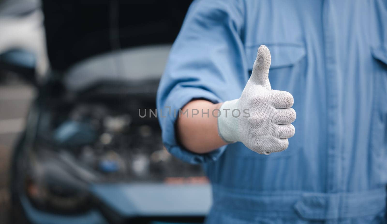 Mechanic in jumpsuit checks car gives thumbs up. Expertise in repair and service ensuring safety and quality. Skilled technician working in automotive industry smiling.