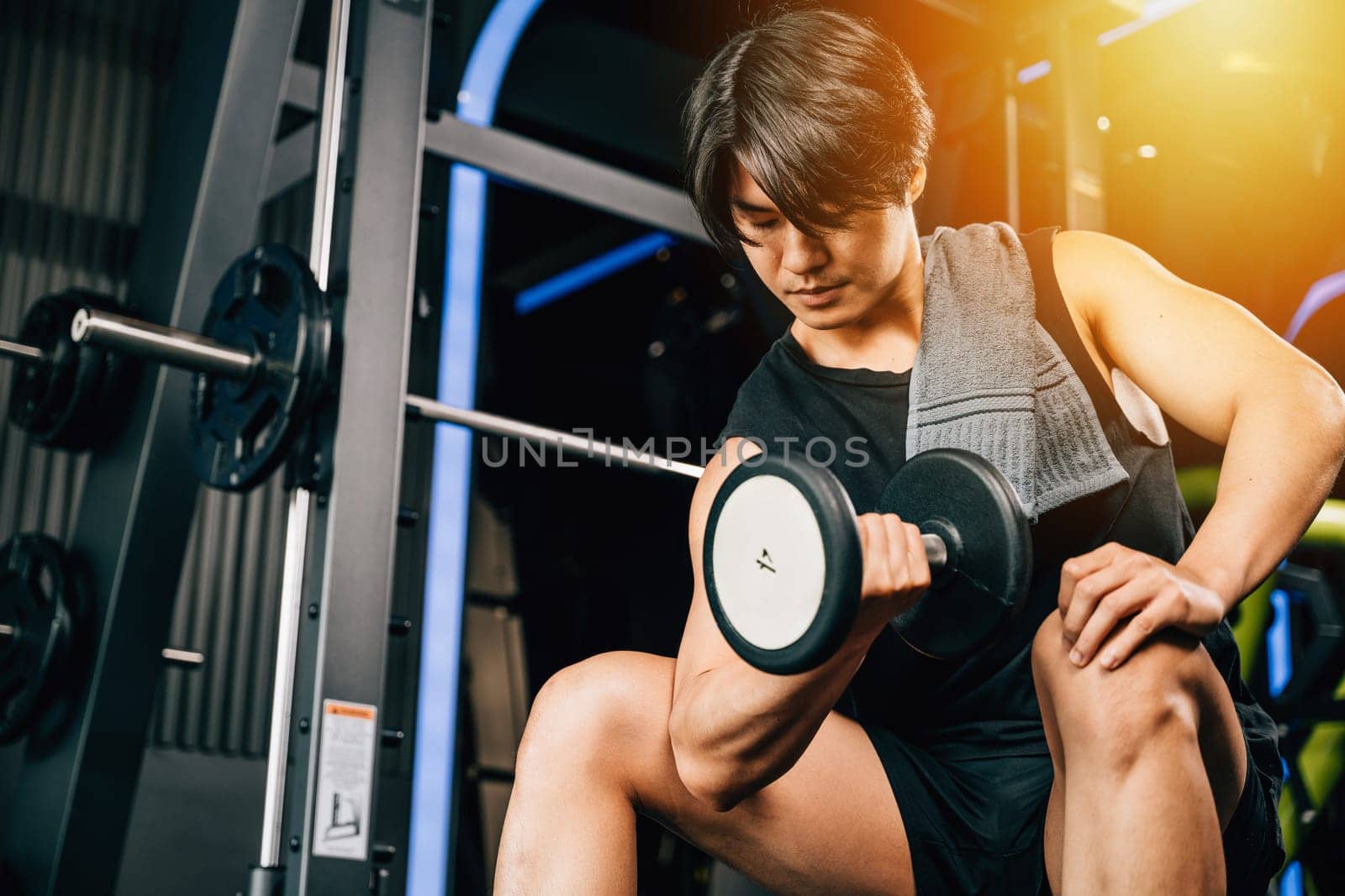 handsome young Asian man sitting on an exercise bench holding a heavy dumbbell by Sorapop