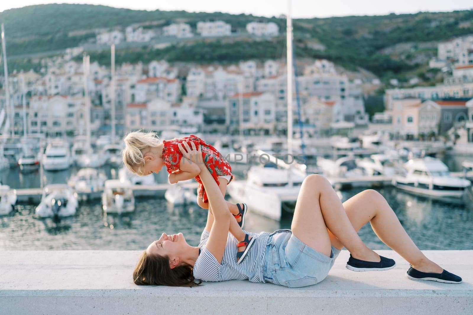 Smiling mother holds a little girl in outstretched arms while lying on the fence of the marina embankment. High quality photo