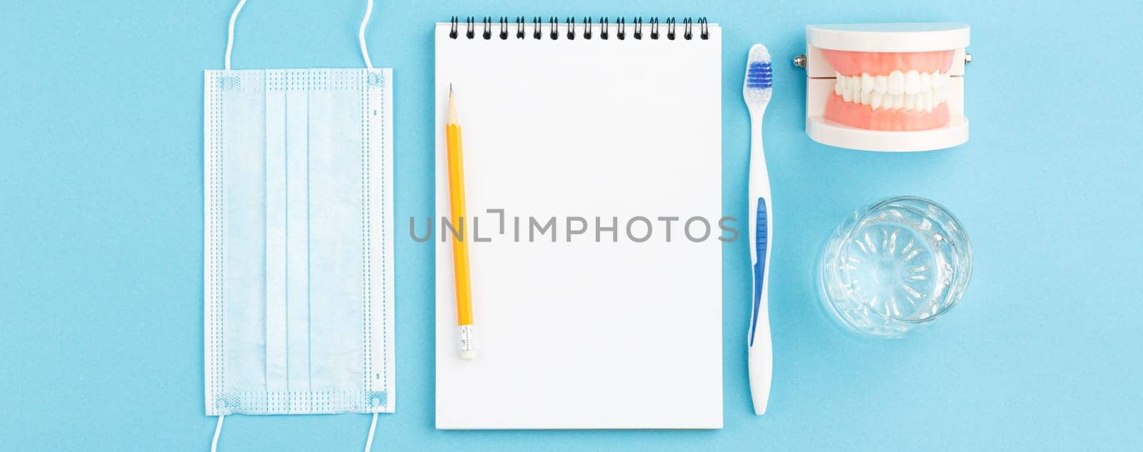 Oral hygiene. Spiral notepad with pencil on blue background. Dental concept. Jaw model with toth brush, glass of water and medical mask on the dentist table. top view.
