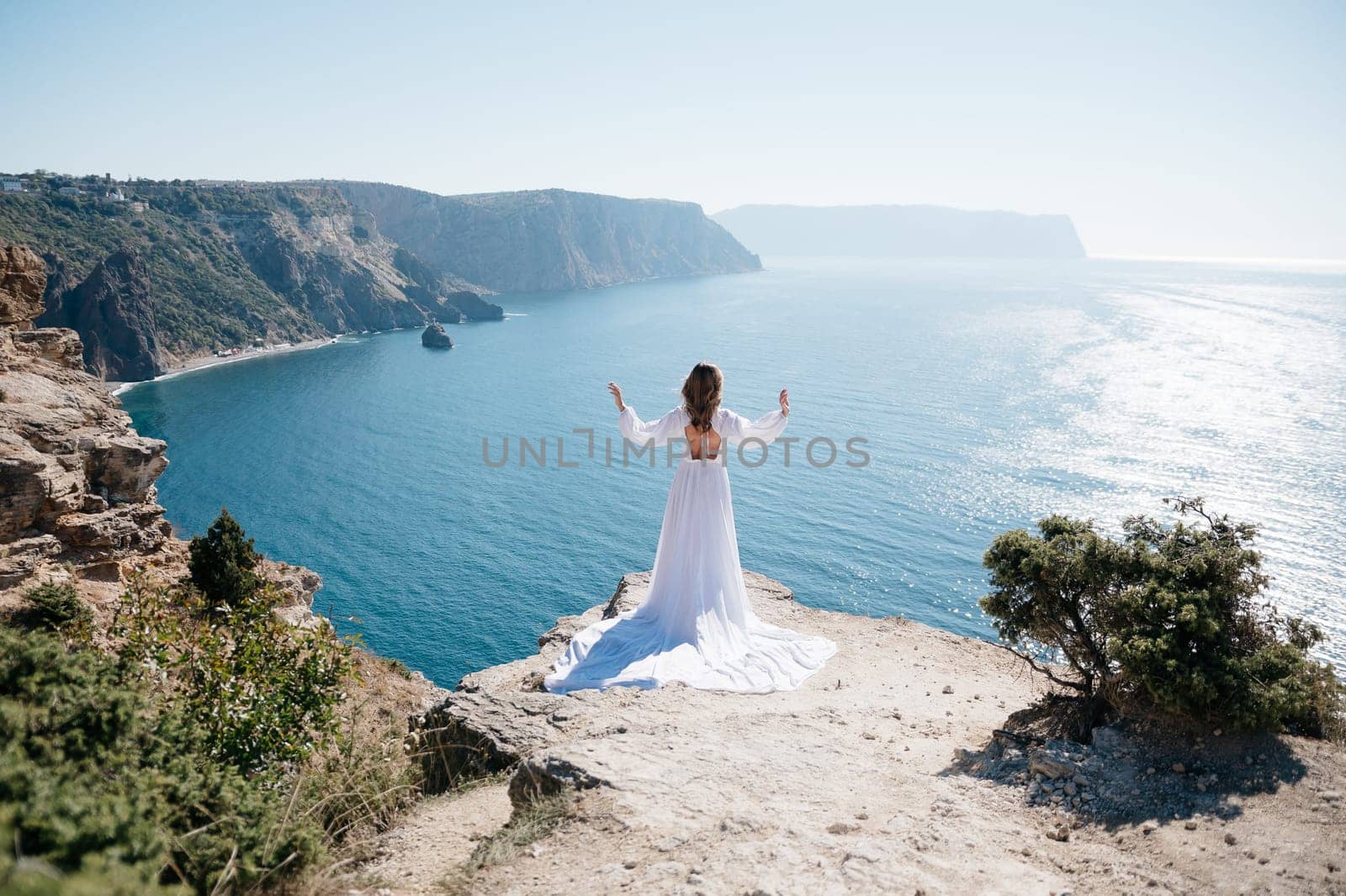 A woman stands on a cliff overlooking the ocean in a white dress. She is looking out at the water and she is in a state of peace and serenity. Concept of calm and tranquility