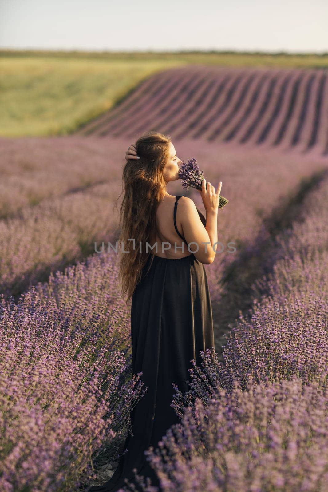 woman stands in a lavender field of purple flowers, holding a bouquet of flowers. The scene is serene and peaceful, with the woman taking a moment to enjoy the beauty of nature. by Matiunina