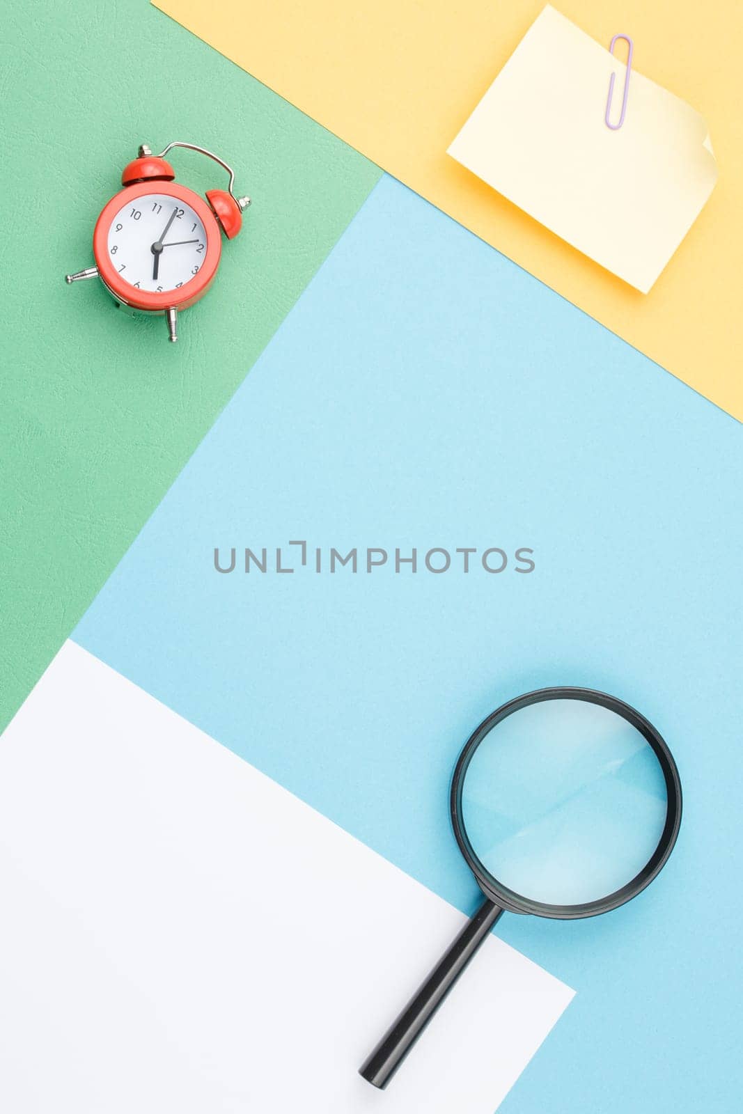 Colored paper background of geometric shapes with magnifying glass, alarm clock, note paper with paper clip and flower in the pot Office composition. Flat lay