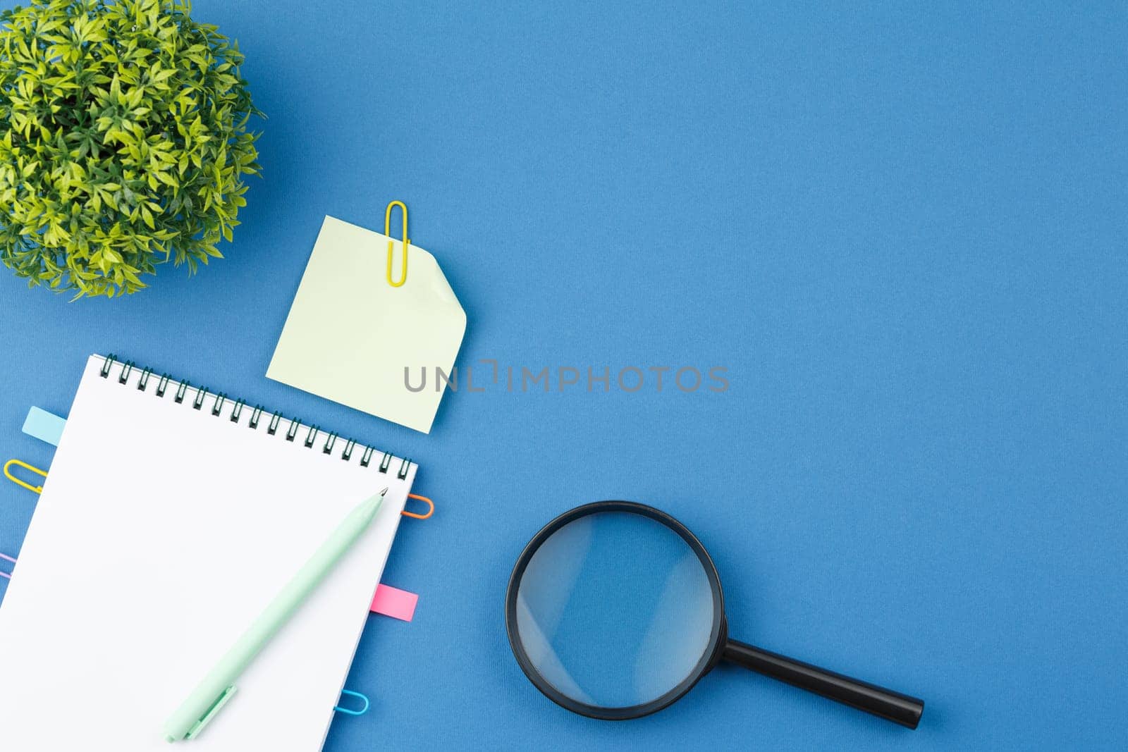Spiral notebook with bookmarks from paper clips, note sheets, pen, magnifying glass and potted plant on blue isolated background. Office concept. Top view.