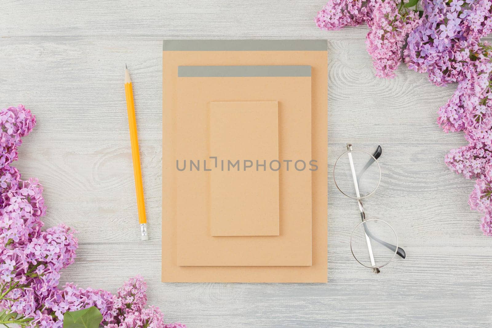 Notebook with pencil, glasses and lilac flowers on a white wooden background. Top view.