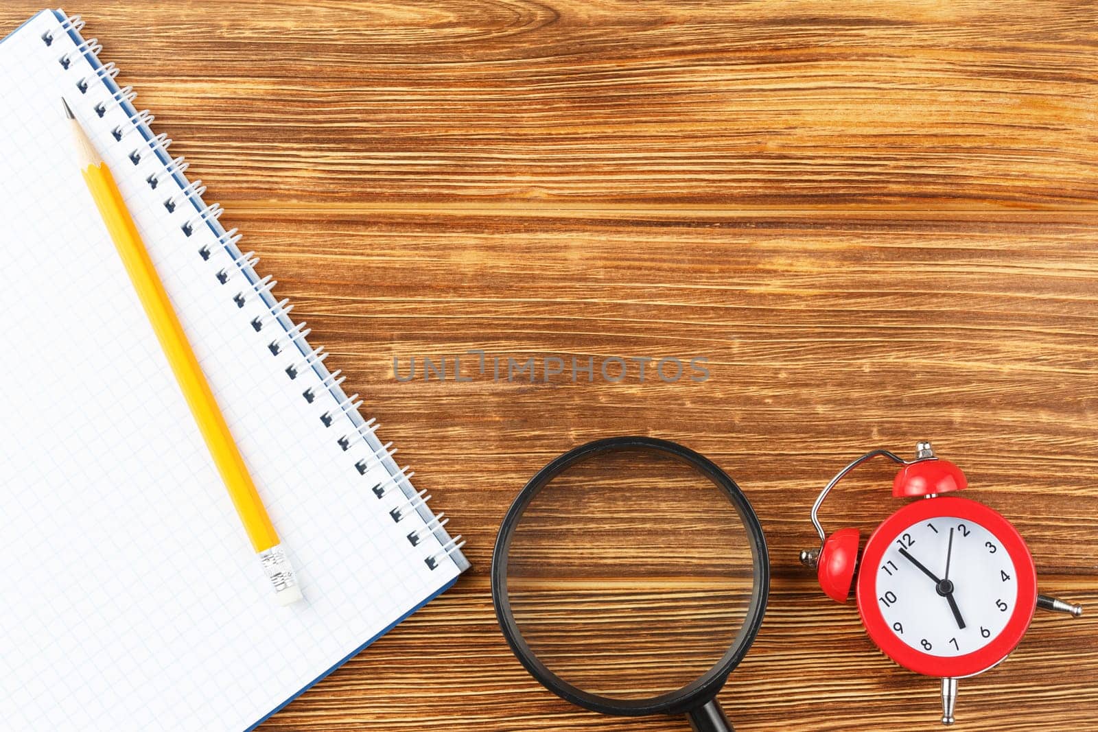 Open spiral notebook with pencil and magnifying glass on wooden background. Study desk with alarm clock. Flat lay. Back to school concept. Top view.