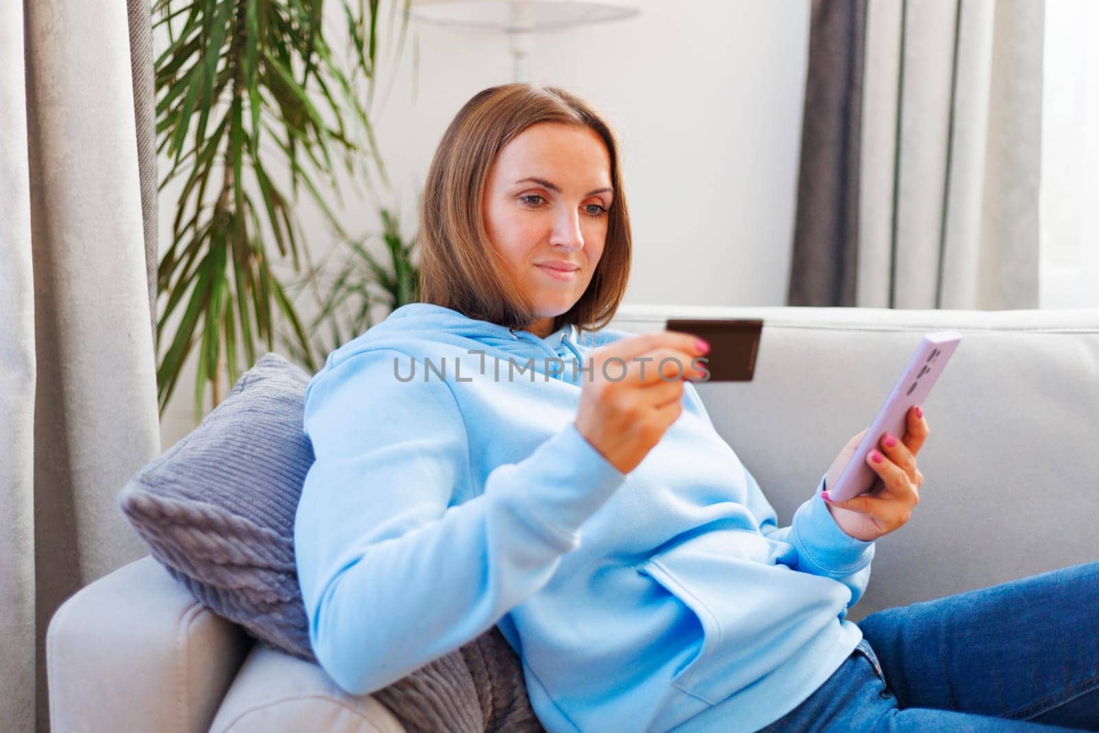 Woman sitting on a couch at home shopping online with a credit card and smartphone. Interior lifestyle portrait. E-commerce and online shopping concept. Design for banner, poster, website header.